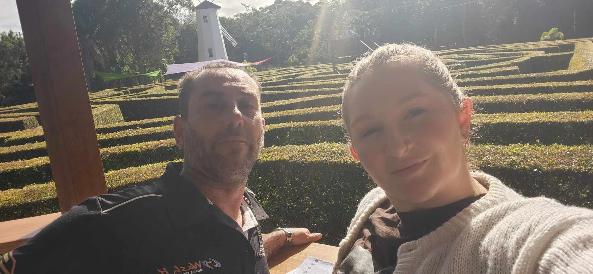 Two people taking a selfie outdoors, with a windmill and terraced garden in the background.