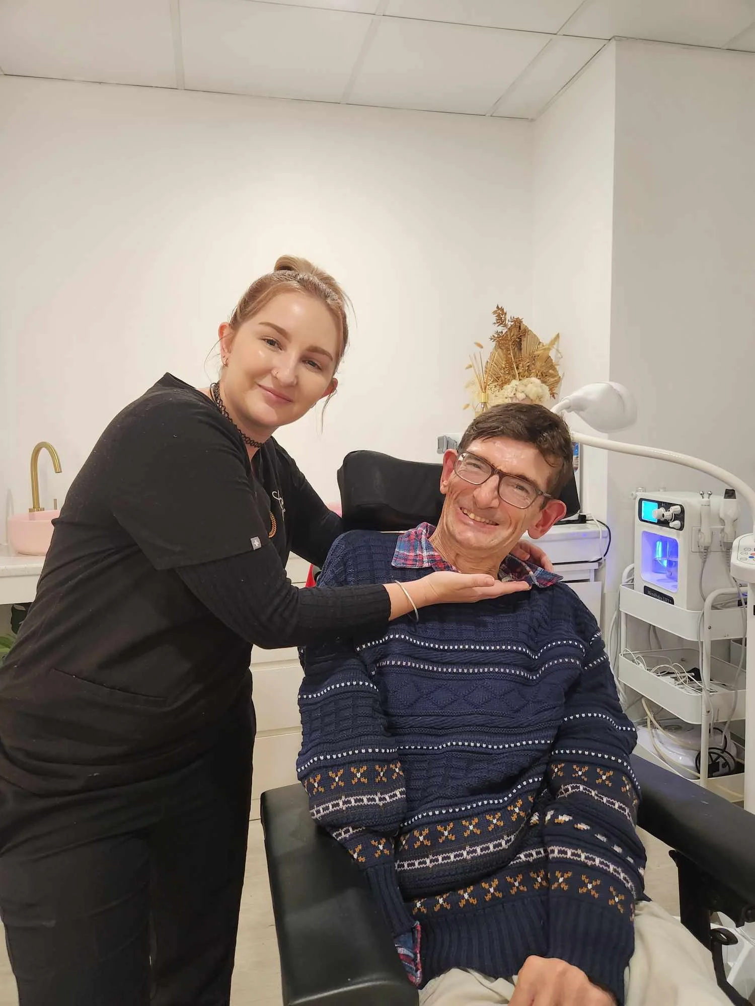 A woman and a man smiling at the camera, the woman is supporting the man's chin with her hand. The man is sitting in a chair in a healthcare or dental office, with medical equipment in the background.
