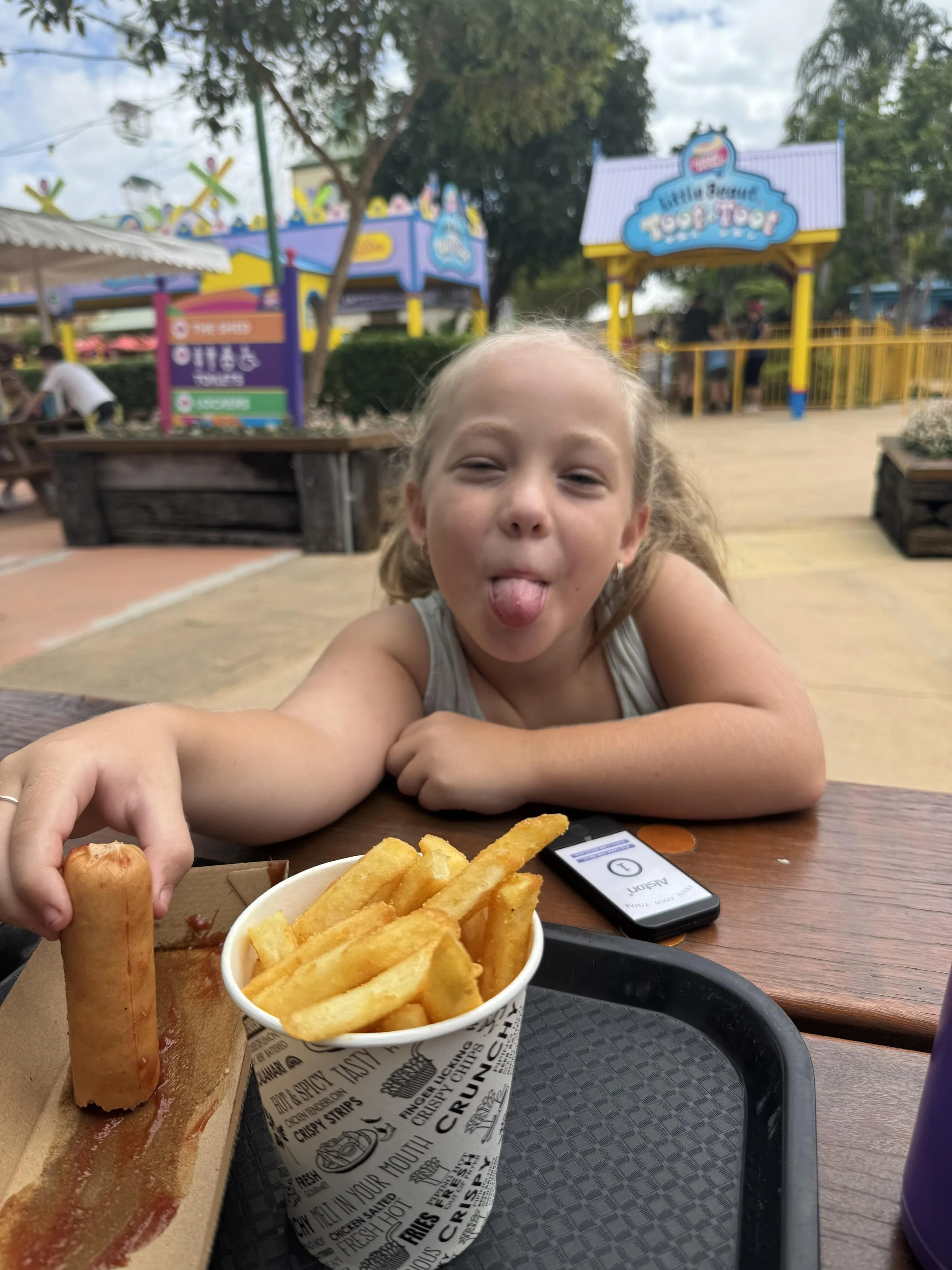 A young girl sticking out her tongue at an amusement park, sitting at a picnic table with a tray of French fries and a sausage hot dog in front of her. In the background, colorful amusement park rides and signs are visible.