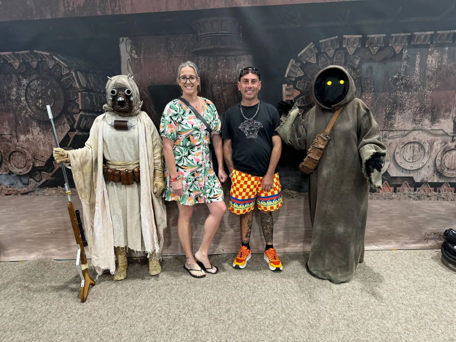 Two people posing with two individuals in costumes from the Star Wars universe, standing in front of a large industrial-themed backdrop.