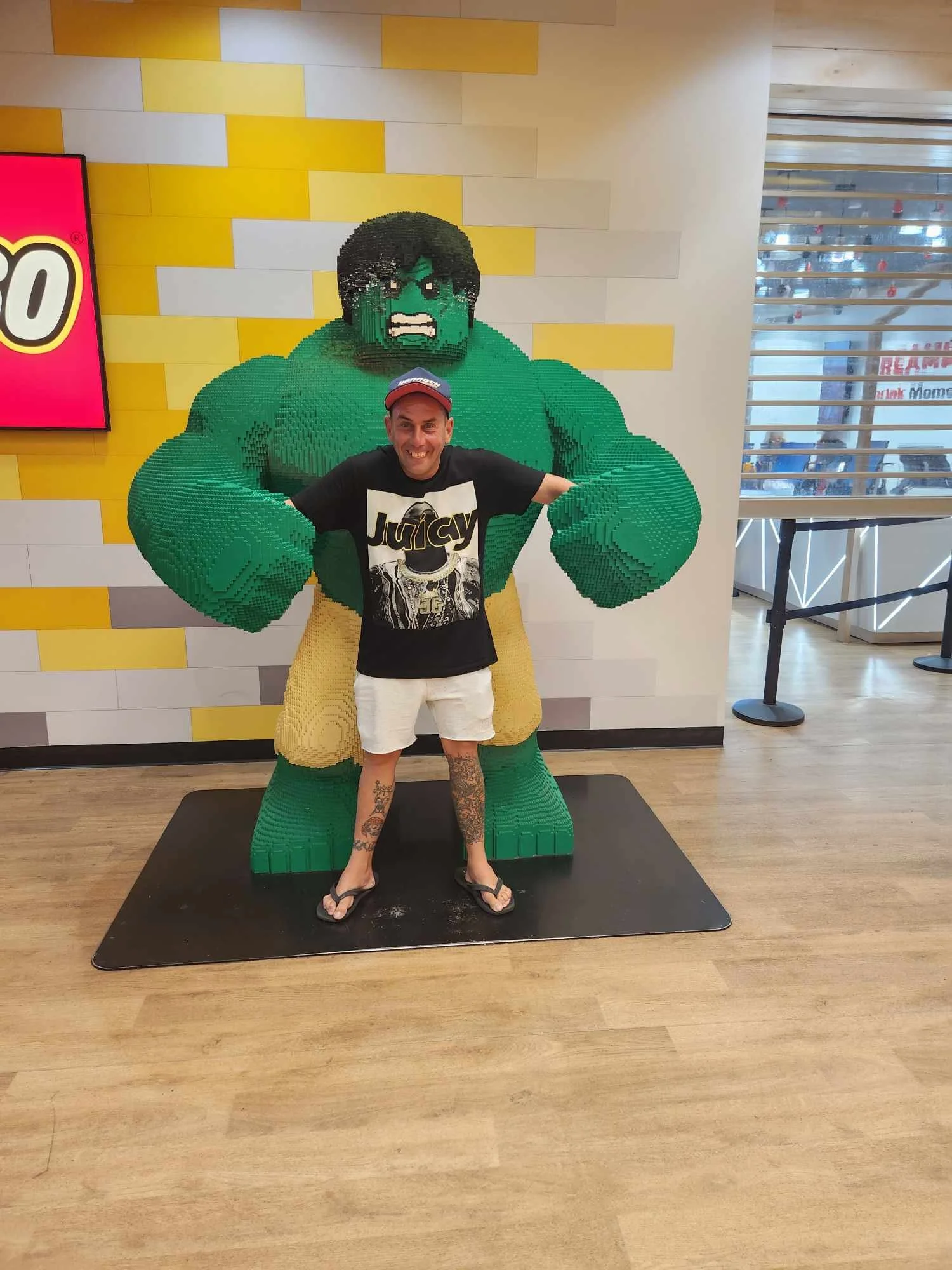 A man standing in front of a large LEGO statue of the Hulk inside a LEGO Store. The man is smiling, wearing a black Juicy t-shirt, white shorts, flip-flops, and a red cap.