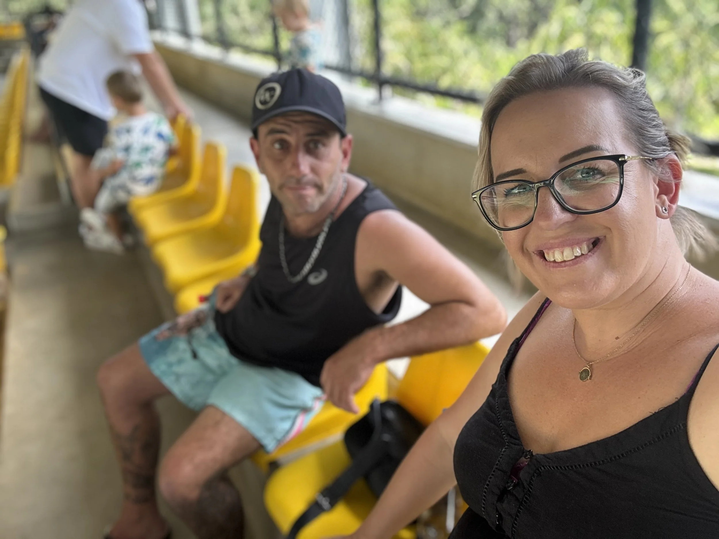 A woman with glasses taking a selfie with a man sitting on yellow bench seats at a zoo exhibit, with children and a woman in the background.