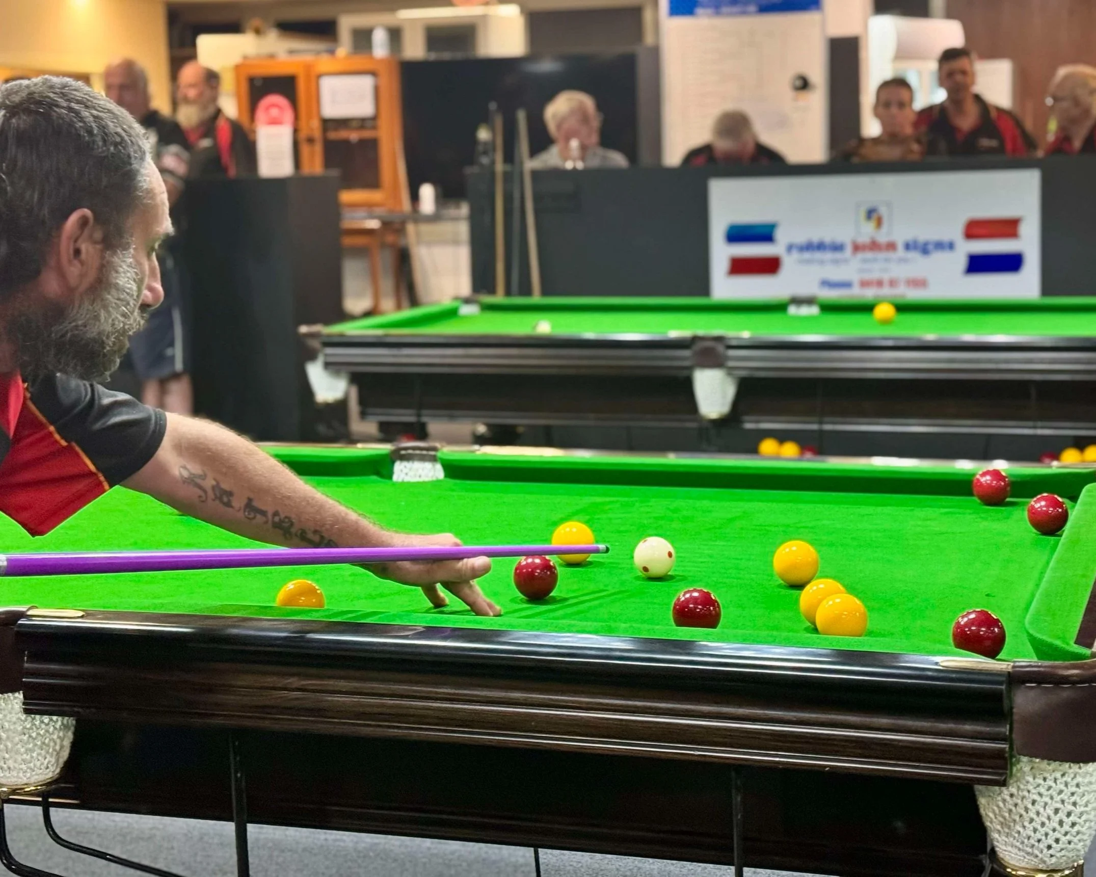 A man playing snooker at a table with yellow, red, and white balls, with onlookers watching in the background.
