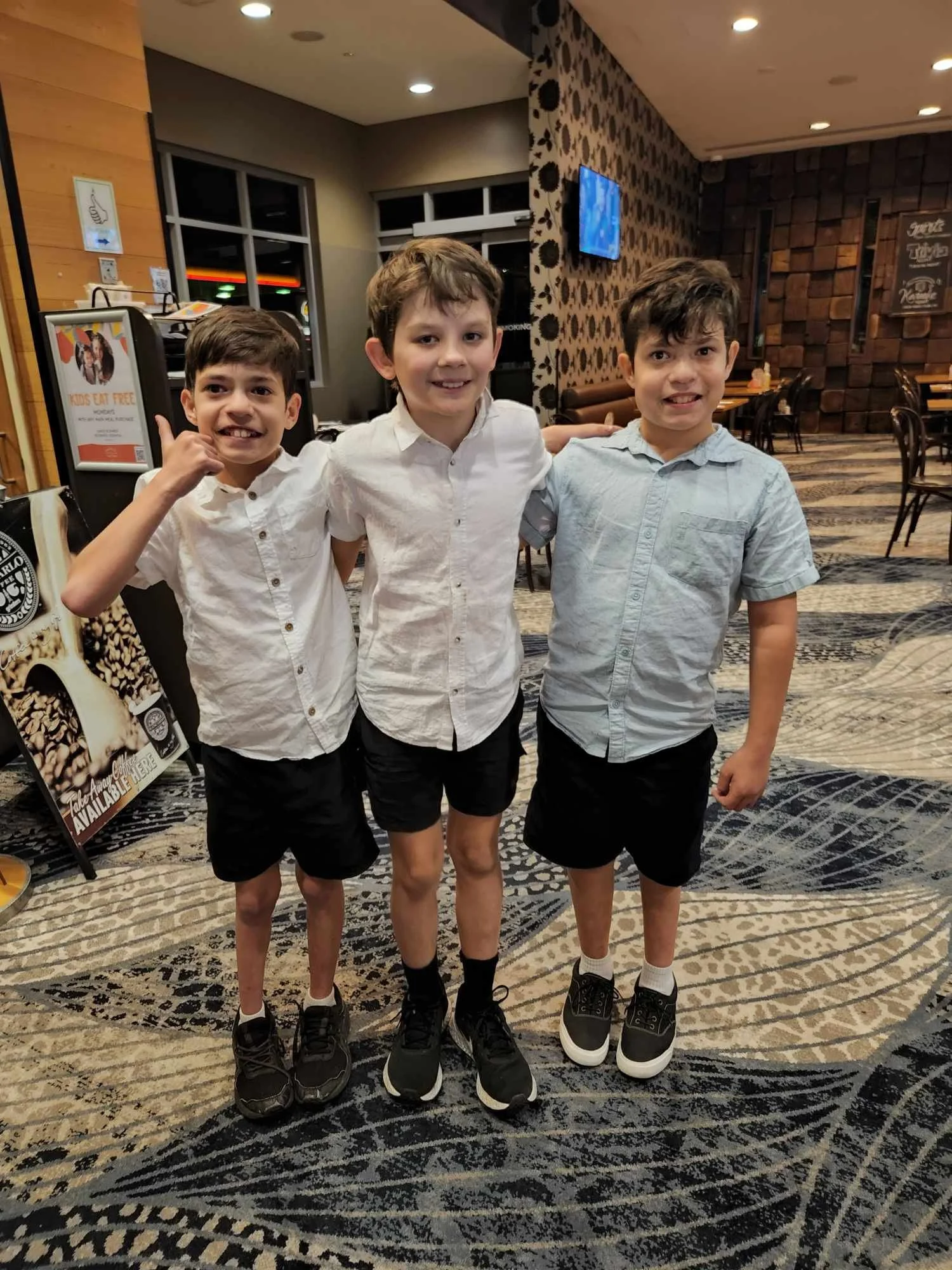 Three young boys in white shirts and black shorts standing together in a restaurant, smiling and posing for the photo.