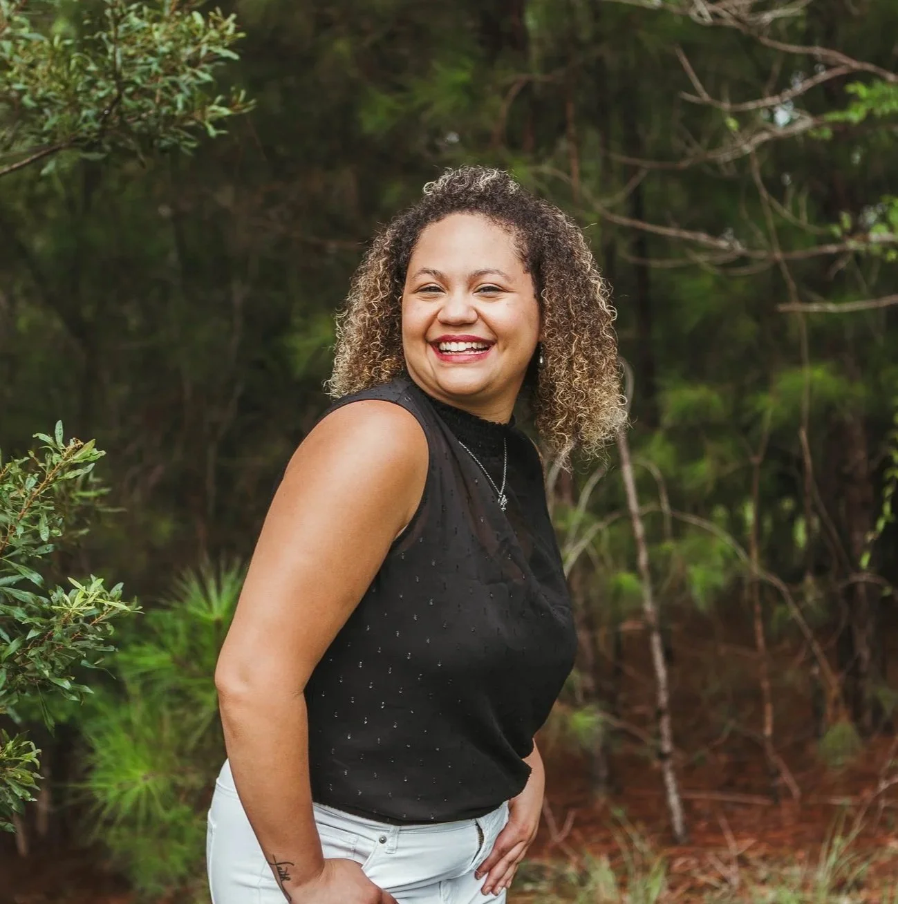 A young woman smiling outdoors in a forested area, wearing a sleeveless black top and white pants.