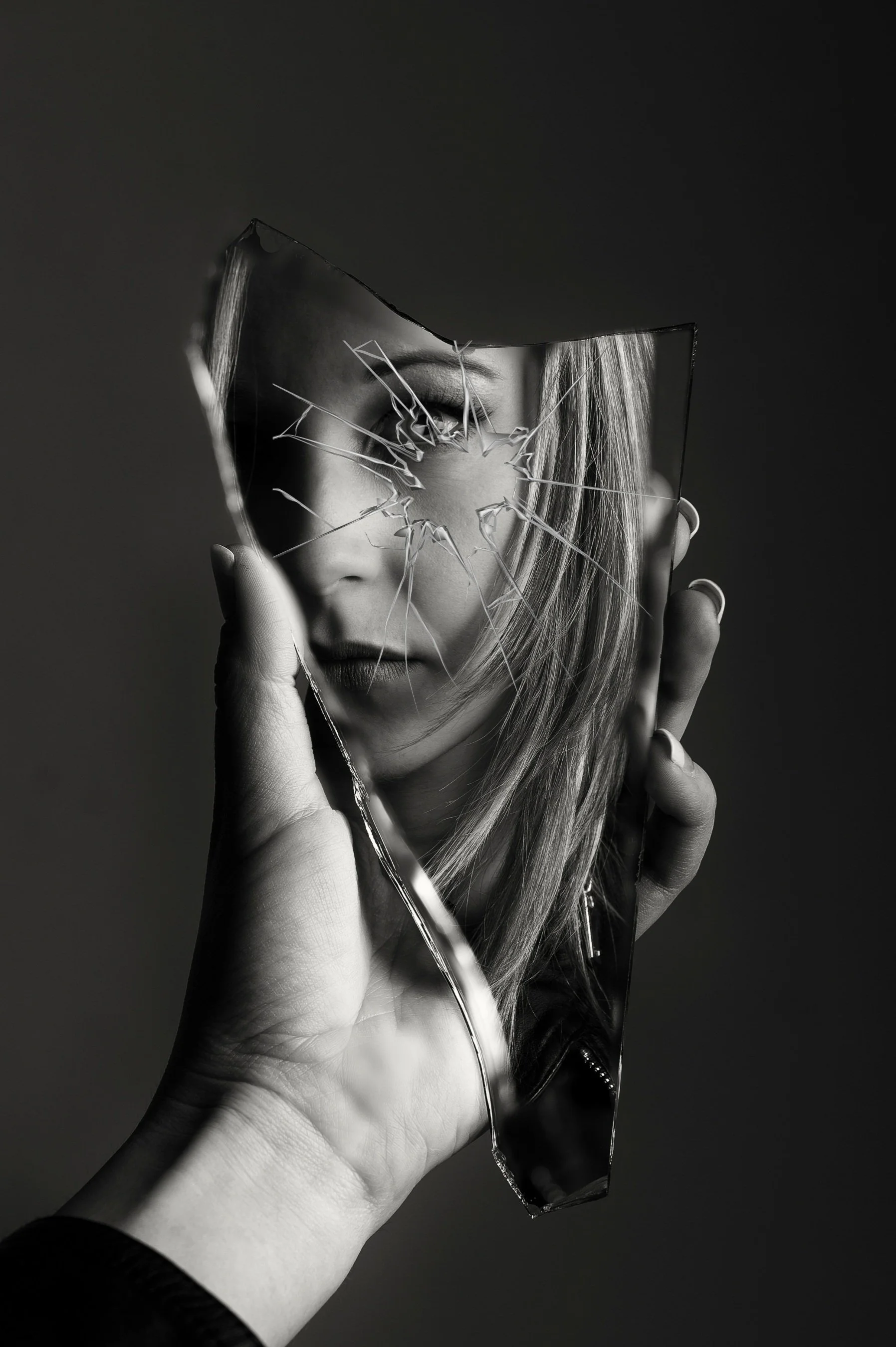 A hand holding a broken mirror reflecting a woman's face with one eye visible, in black and white.