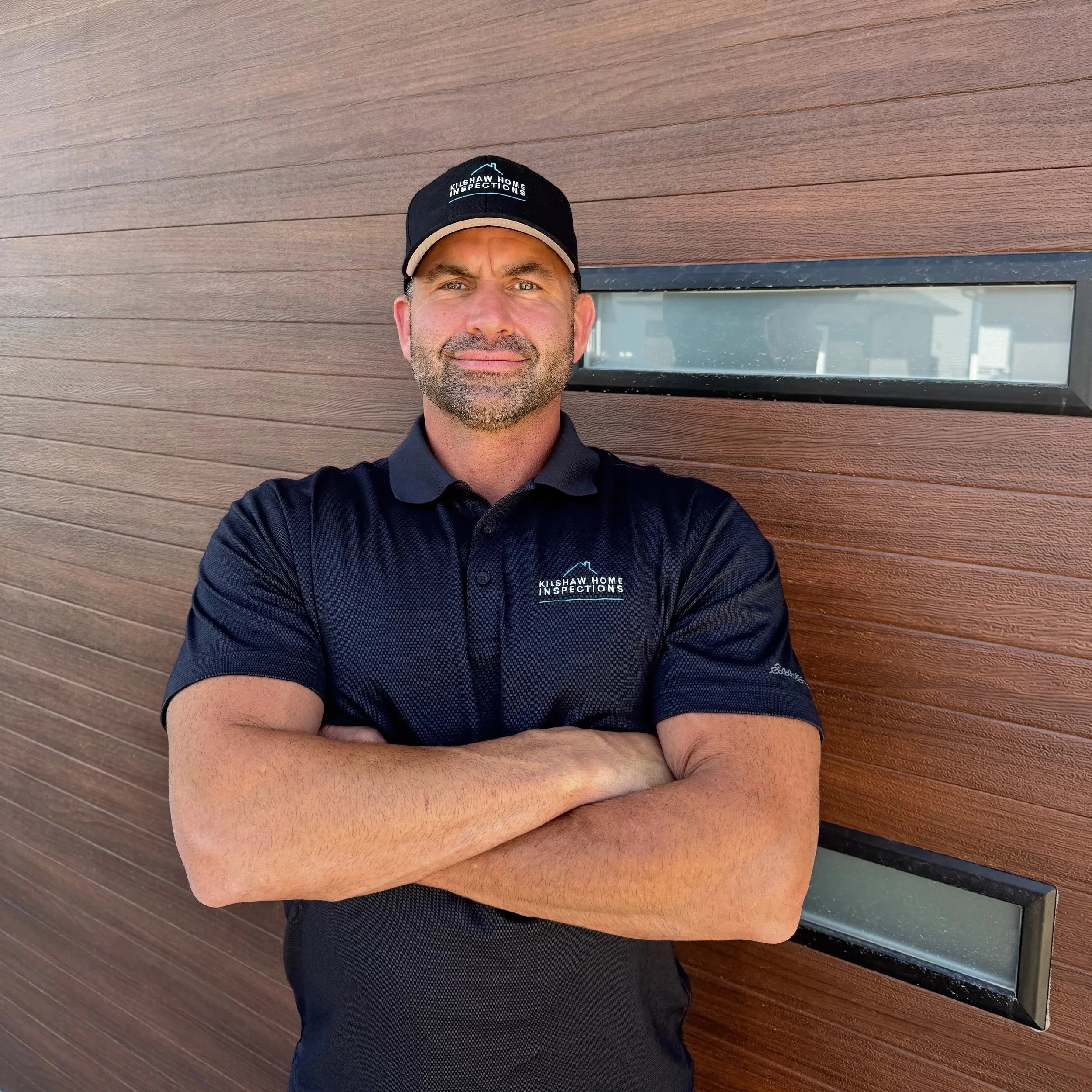A man wearing a black KilShaw Home Inspections polo shirt and cap stands with arms crossed in front of a brown wooden wall with two horizontal windows.