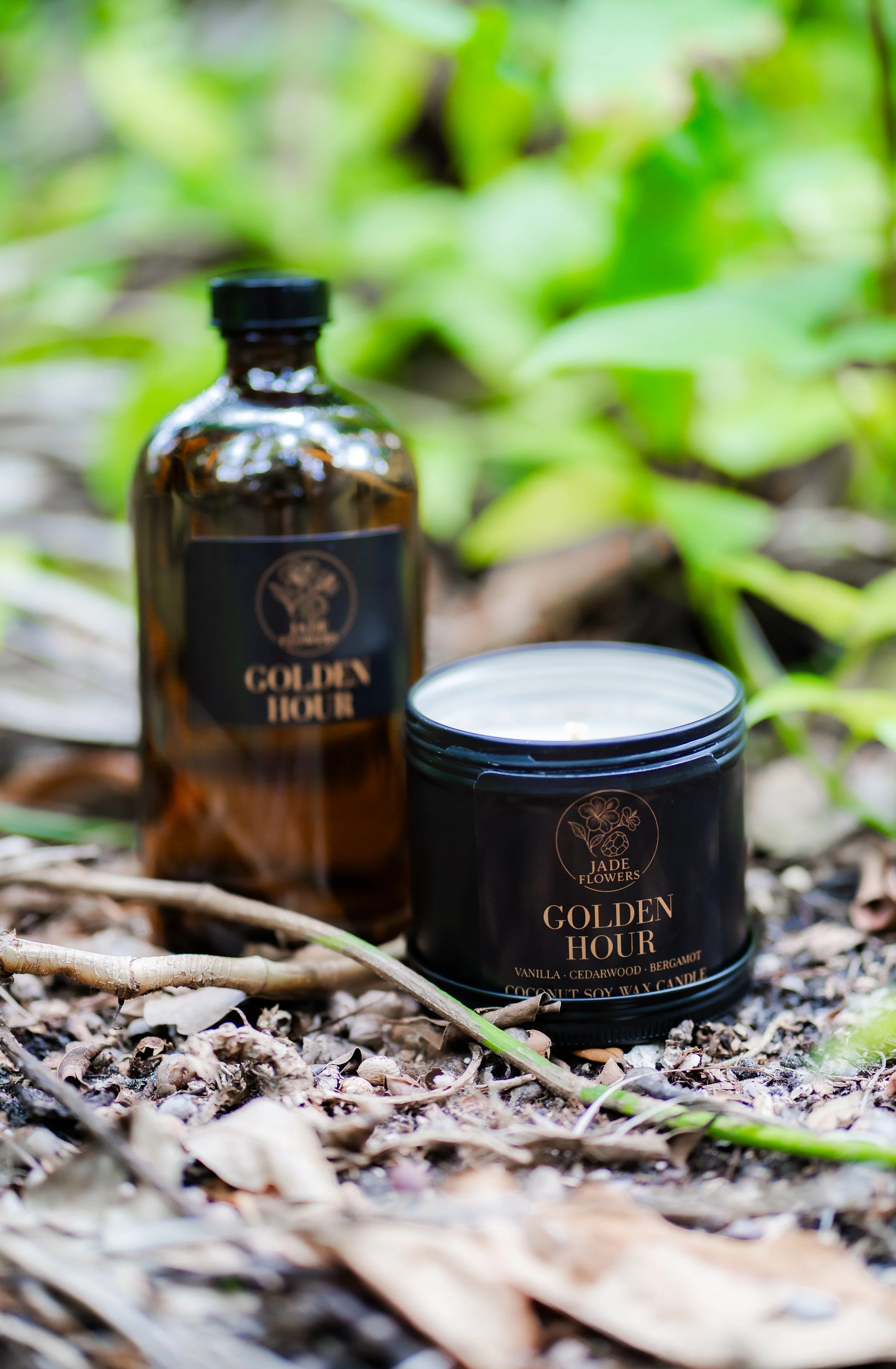 A brown glass bottle and a black candle tin labeled 'Golden Hour' with scents of vanilla, cedarwood, bergamot, placed on the ground among dry leaves and twigs in a natural outdoor setting with green foliage background.