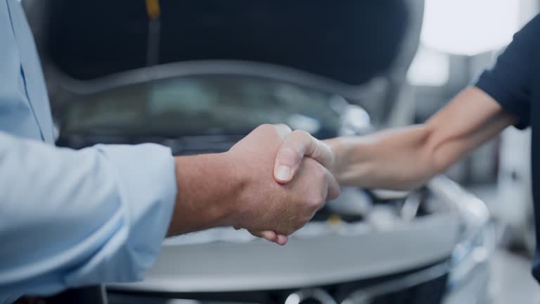 A close-up of a handshake between two people in front of a car with an open hood, likely at an auto repair shop or service station.