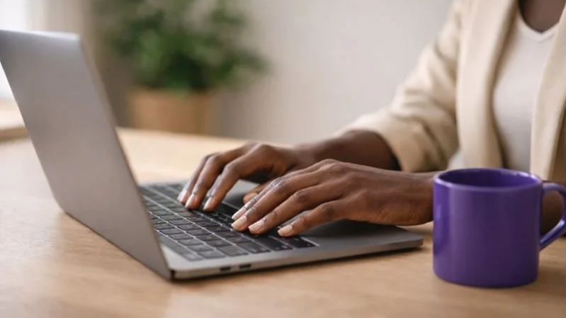 Person typing on a laptop with a purple mug on a wooden table.