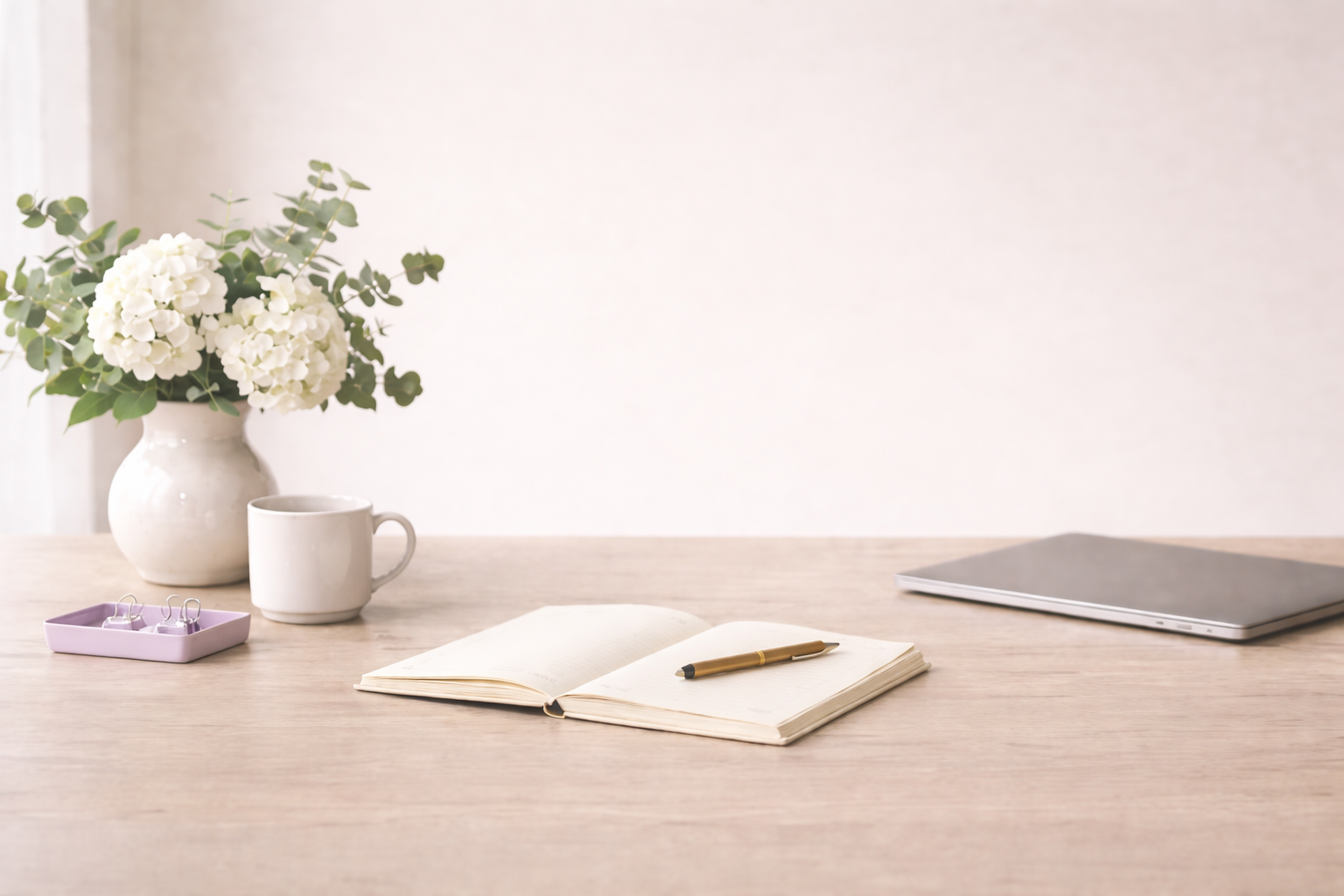 Simple desk with a white vase of white flowers, a white mug, a purple tray with paper clips, an open notebook with a pen, and a closed laptop on a wooden surface.