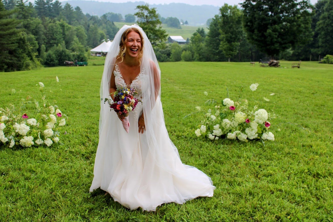 A smiling bride in a white wedding dress with a veil, holding a bouquet, standing on a lush green lawn with white and pink flower arrangements, background of trees, hills, and farm buildings.