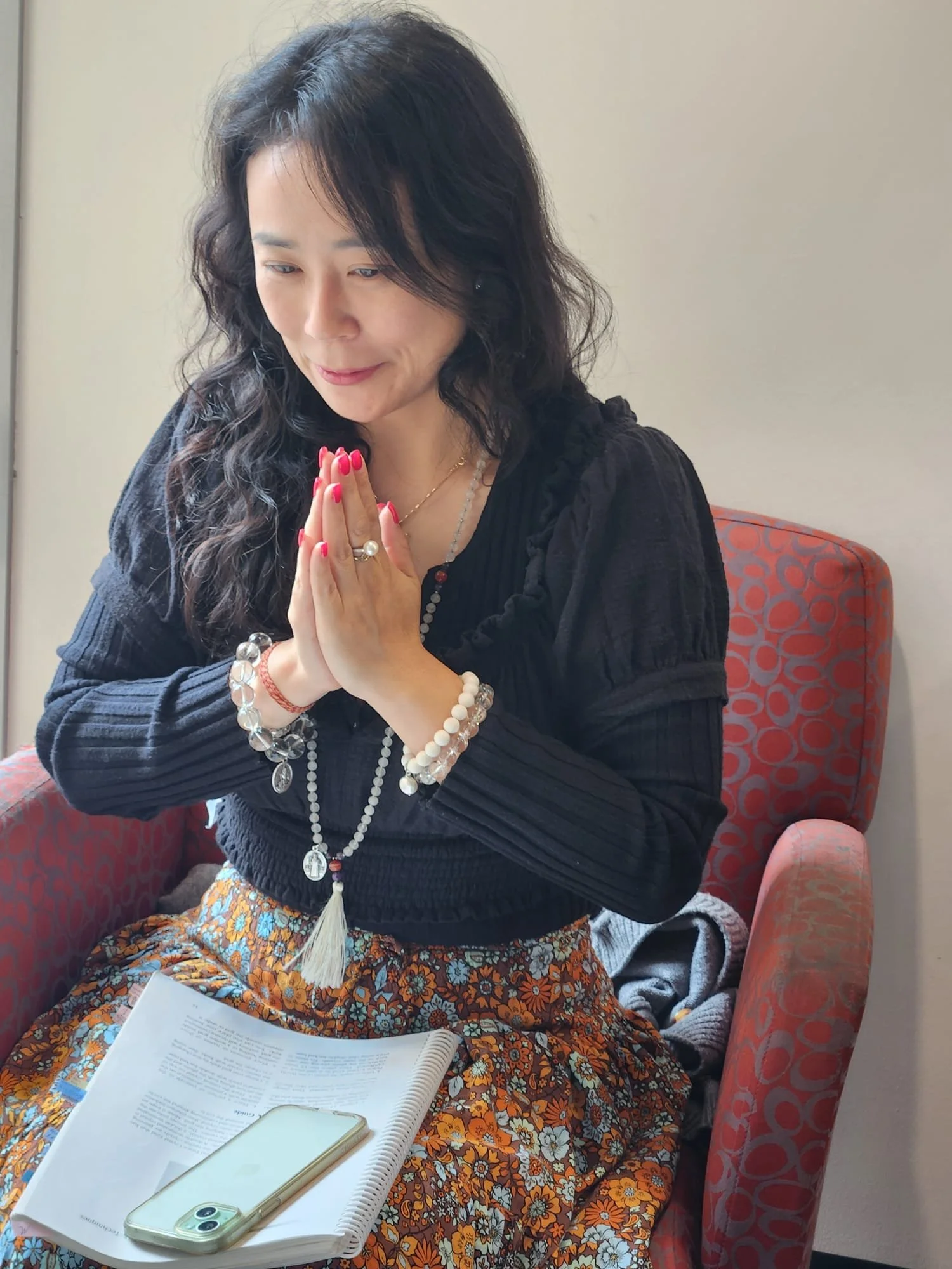A woman with black, wavy hair, wearing a black blouse and floral skirt, sitting on a red patterned chair with her hands together in a prayer position, and looking down.