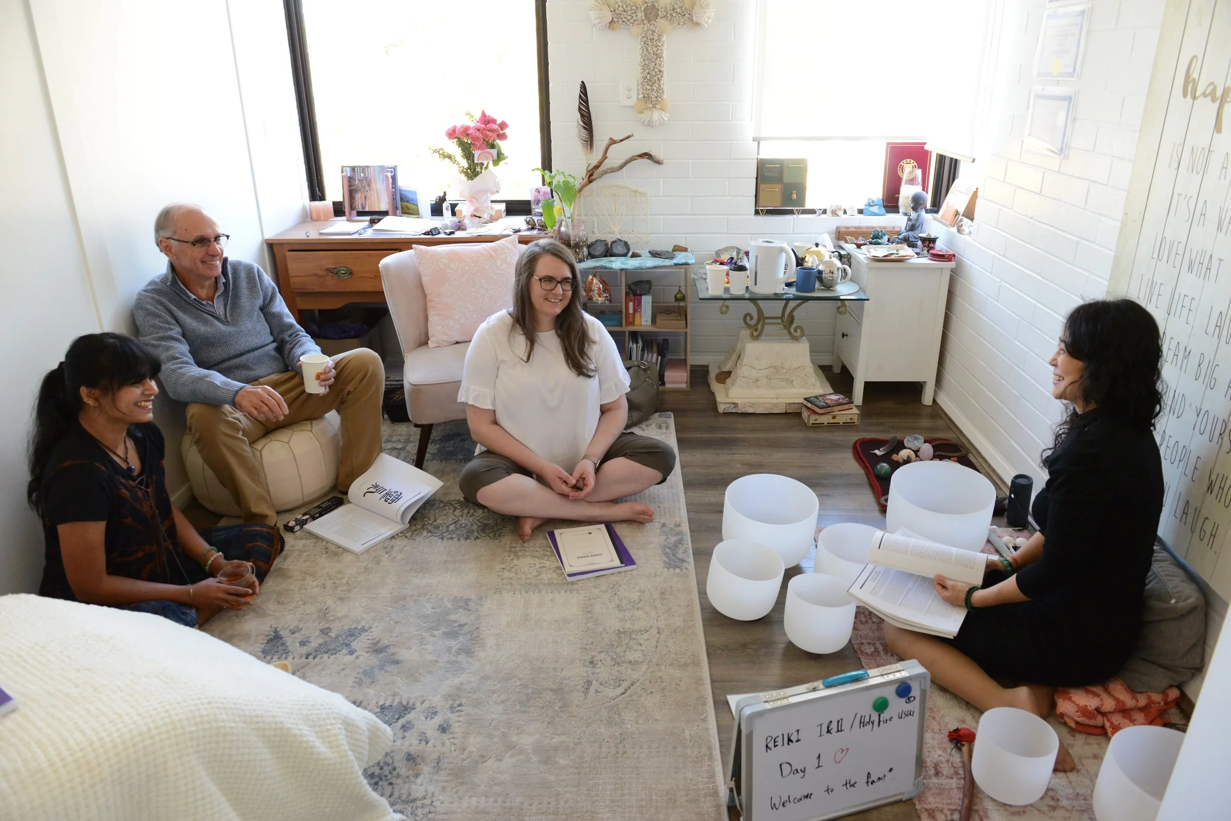 A group of five people sitting in a bright office or living room, engaged in conversation. They are smiling and appear to be having a relaxed meeting. The room has large windows, decorative items, and a small table with books, cups, and a teapot.