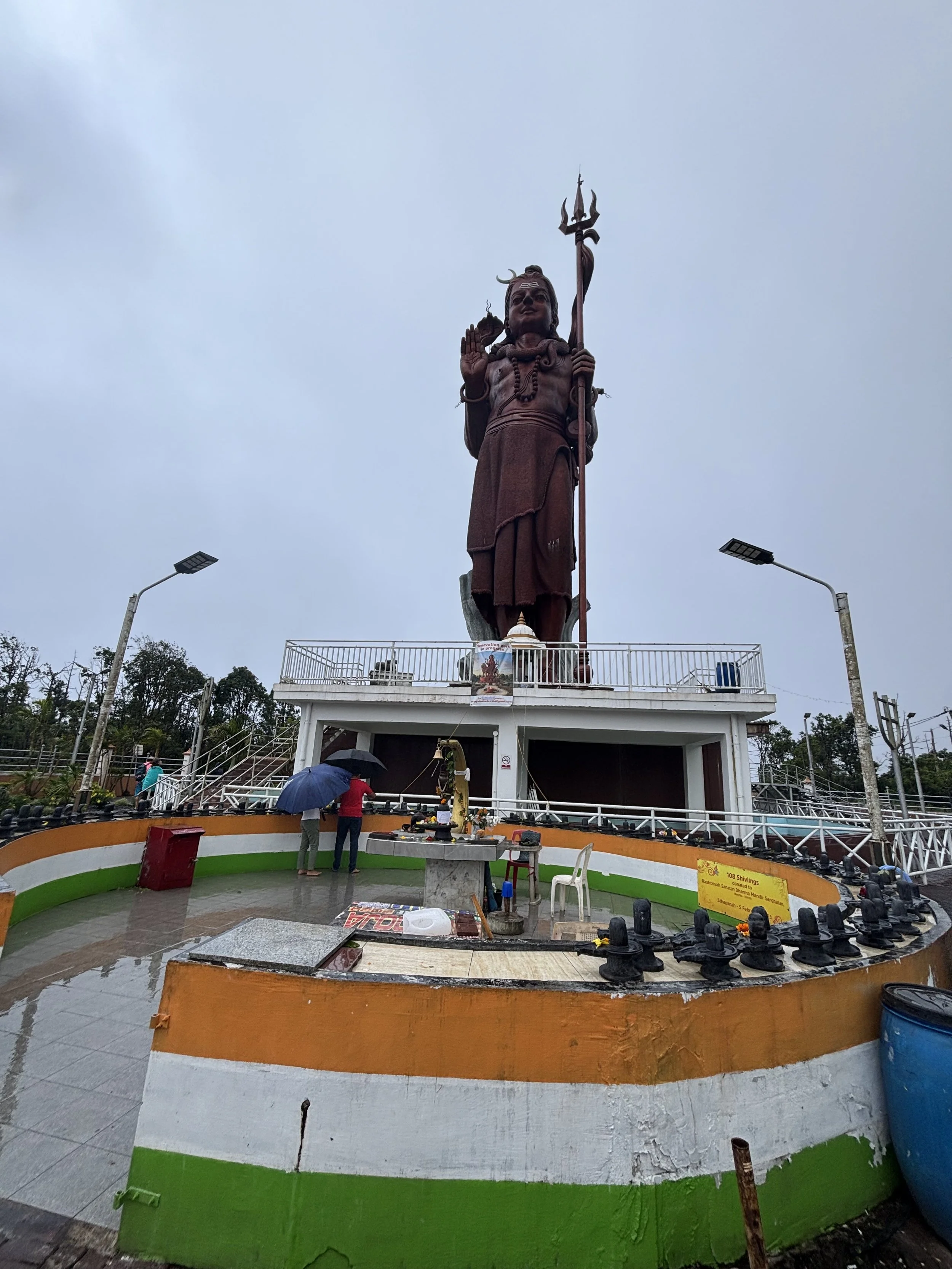 A tall statue of a Hindu god holding a trident, located on a decorated, circular platform painted with the Indian flag colors, with people cleaning the area on a rainy day.
