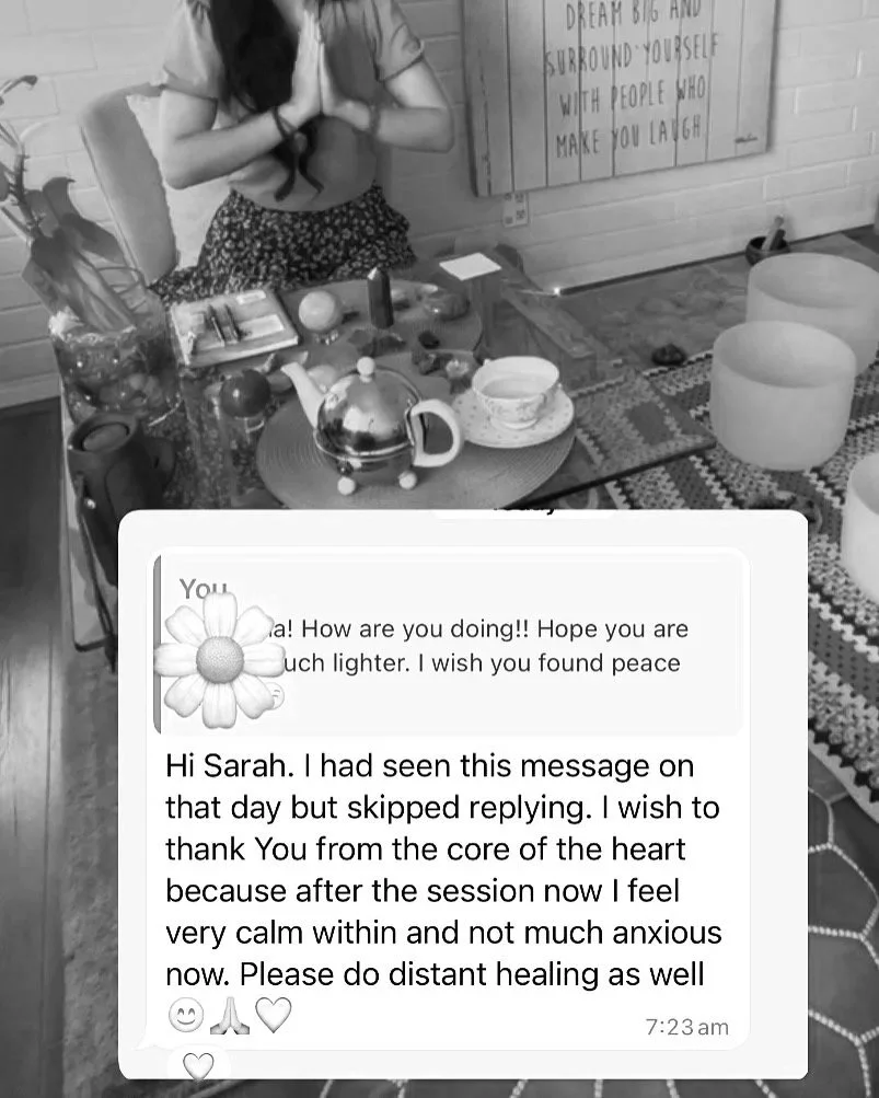 A woman practicing meditation in a room with a table decorated with various crystals, a teapot, a cup of tea, and singing bowls. There are plants and inspirational wall art in the background.