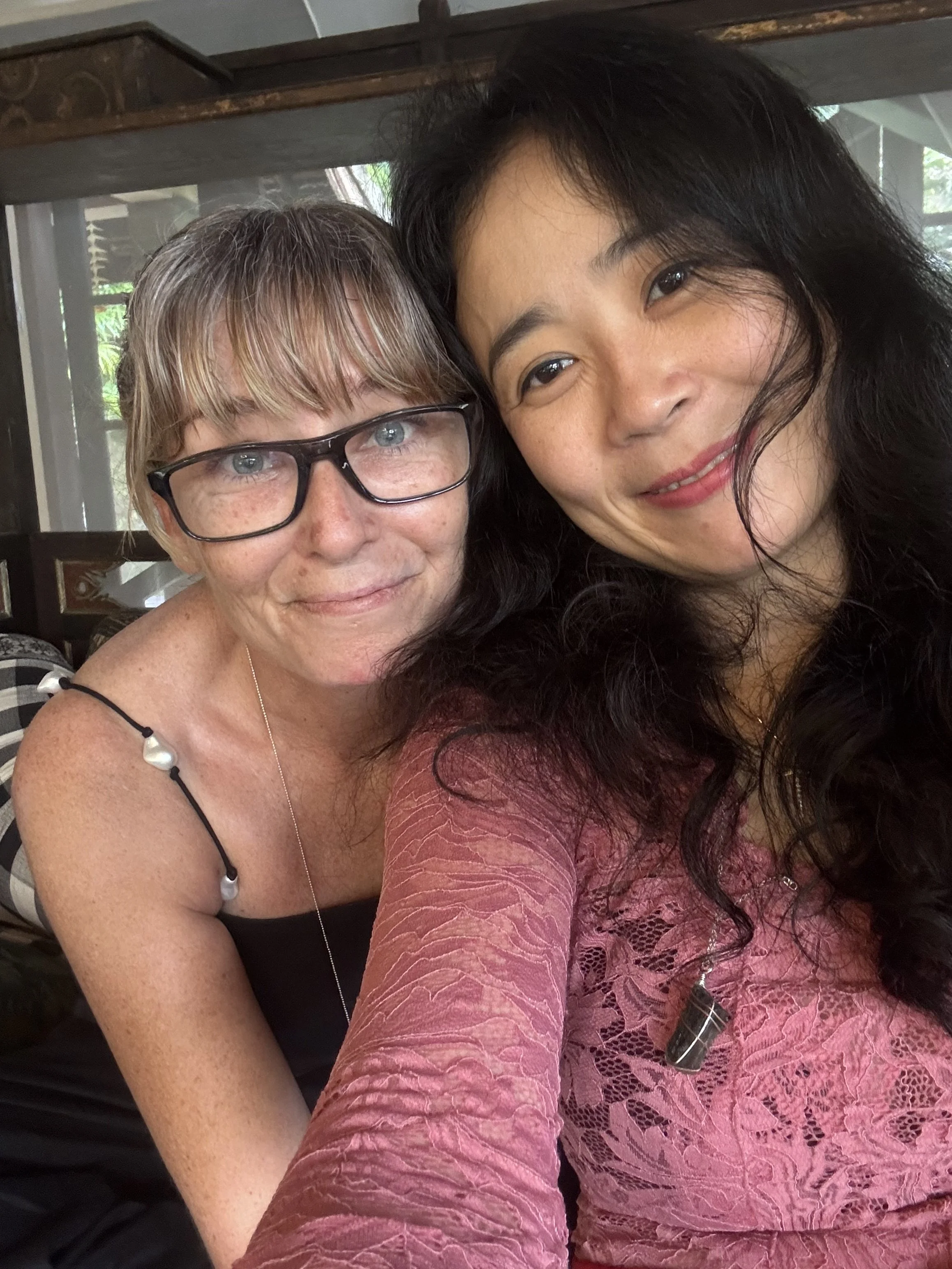Two women taking a close-up selfie together, smiling. One has black hair and the other has blonde hair with glasses. They are inside a house with a window and greenery outside visible in the background.