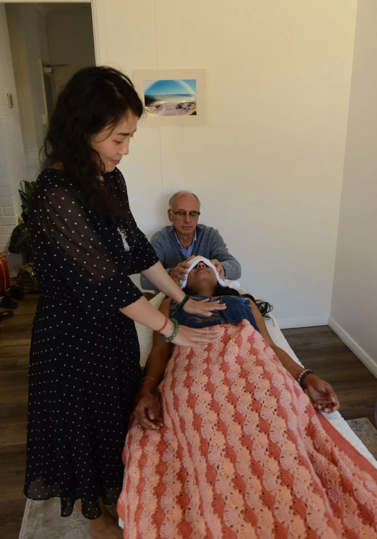 A woman is lying on a massage table, covered with an orange and pink crocheted blanket. Two people are attending to her; a woman on the left gently holds her hand, and an older man behind her supports her head with a towel. The scene takes place in a