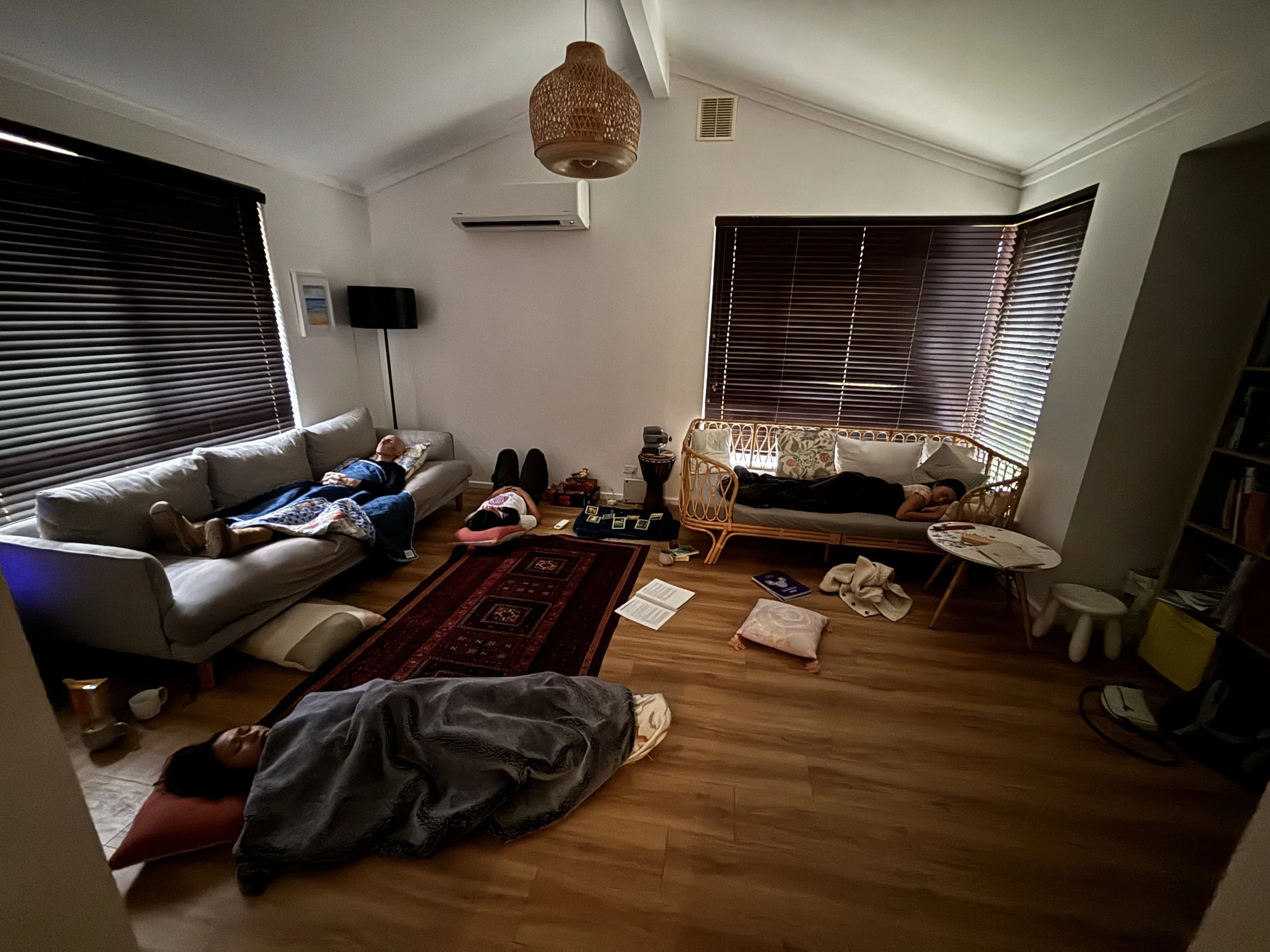 A living room with two people laying on the floor, one on a couch and the other on a blanket, with various furniture and items scattered around, including a rug, a small table, and window blinds.