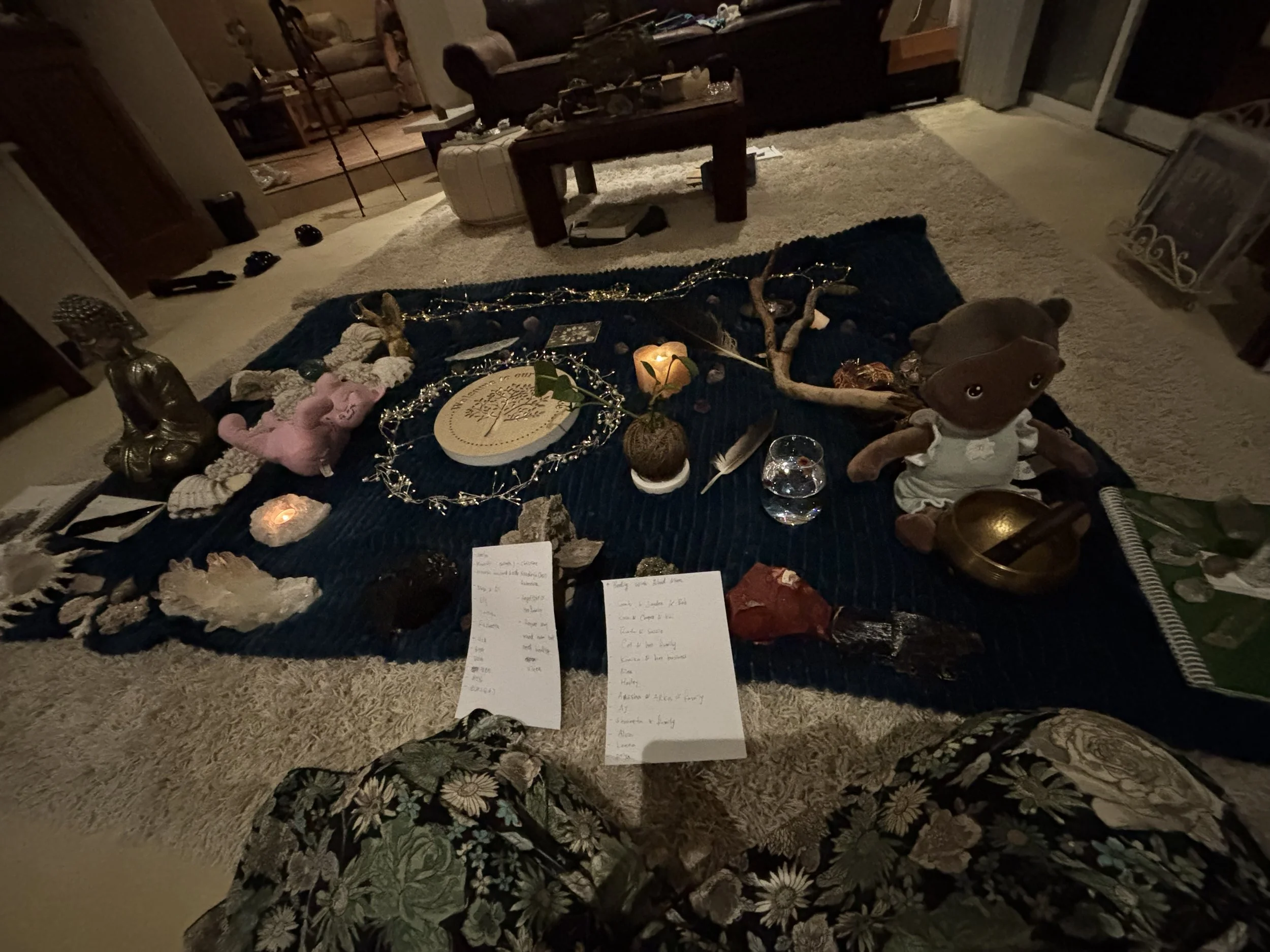 Living room with a table covered in various objects, including candles, crochet items, and handwritten notes; a cozy space with carpeted floors and furniture in the background.