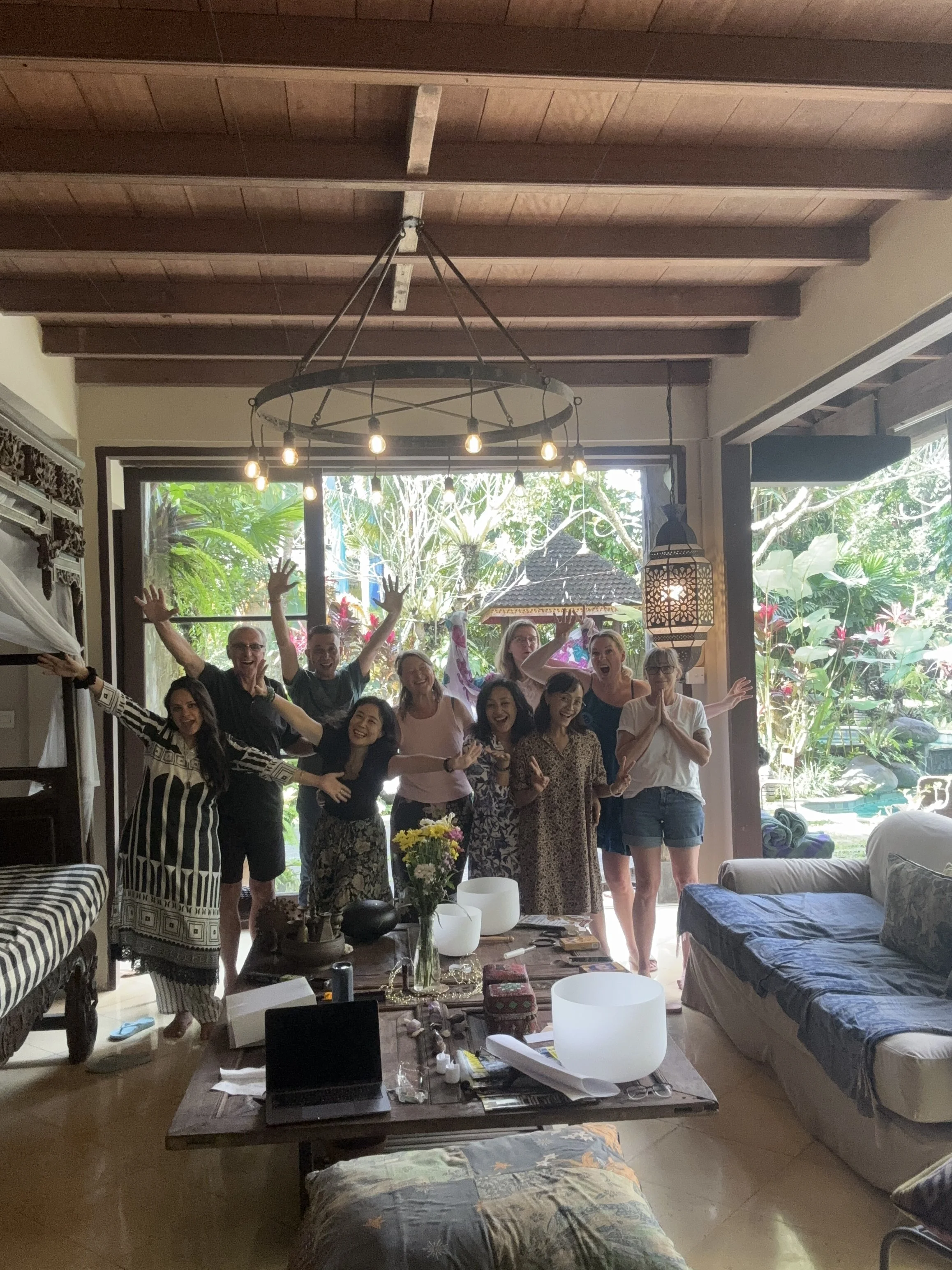 A group of people gathered in a living room, smiling and waving at the camera, with plants and furniture visible in the background.