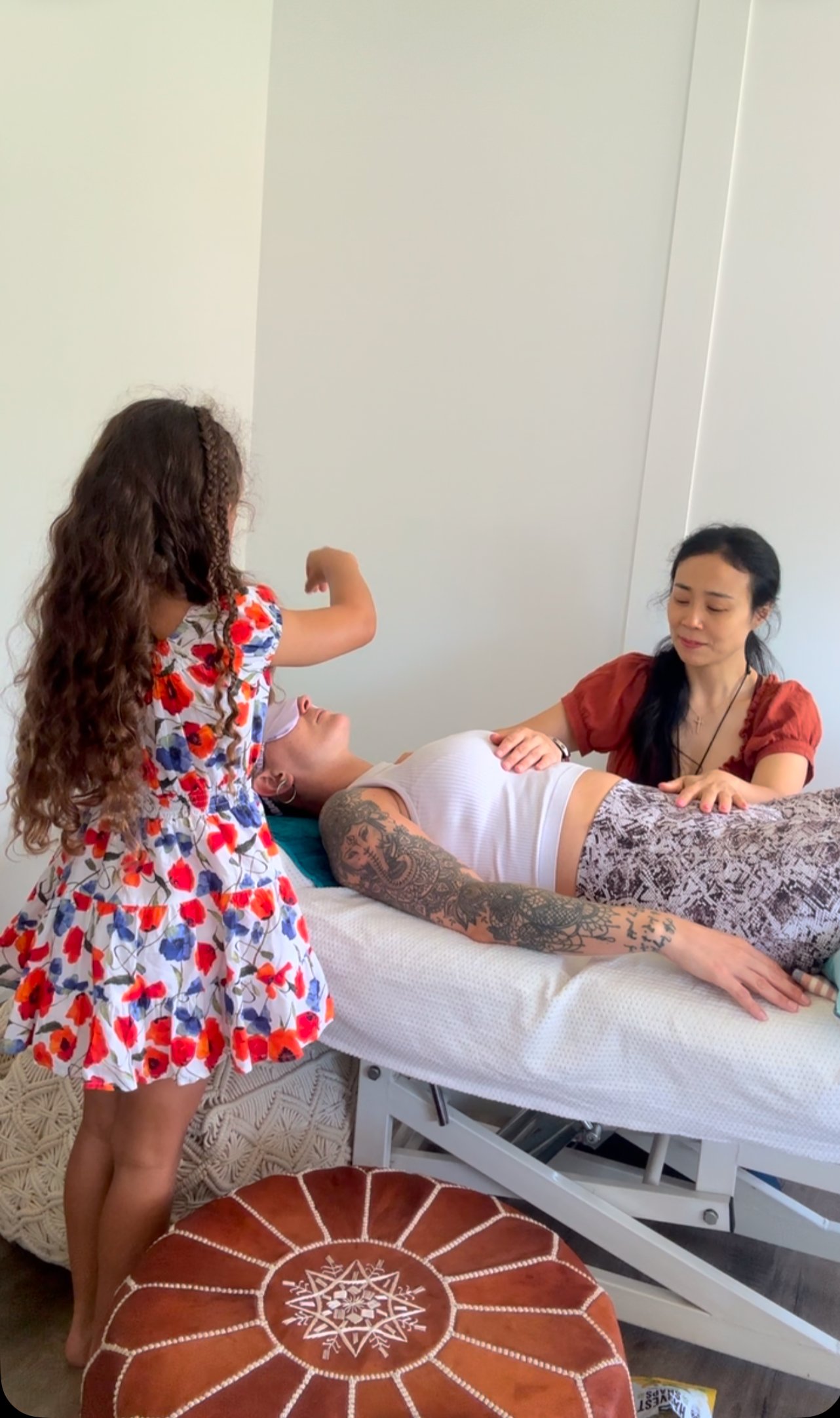 A woman with tattoos lying on a medical bed and a woman with black hair in a red shirt sitting next to her, with a young girl with curly hair and a floral dress standing nearby. The girl is reaching towards the woman on the bed.