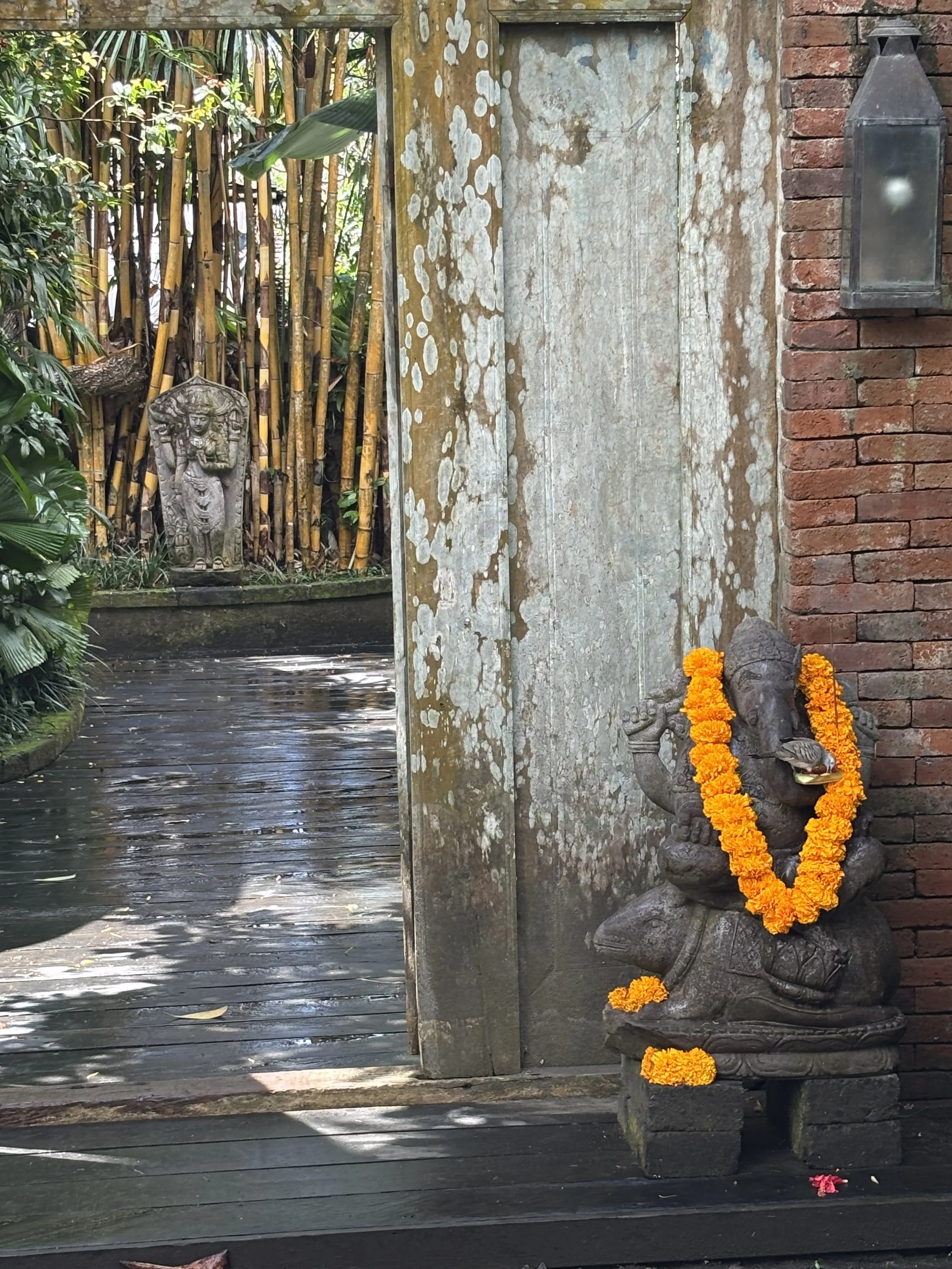 A stone sculpture of a sitting figure adorned with an orange marigold garland, placed against a brick wall. A small red flower is on the ground nearby, and a nearby stone carving of a deity or figure is also decorated with a marigold garland. The sce