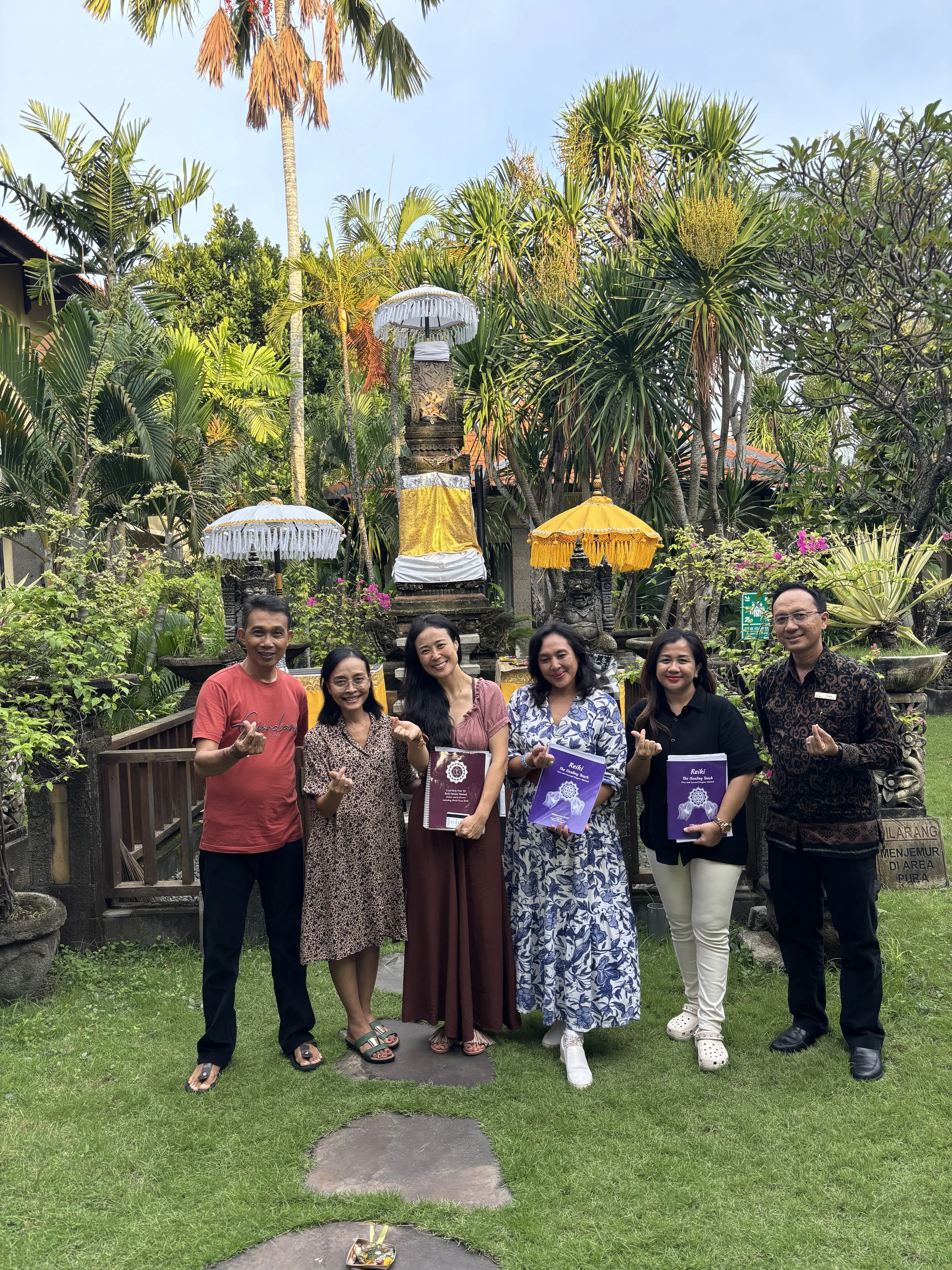 Group of six people standing outdoors in a garden with tropical plants and statues, holding books and making a gesture with their hands, smiling at the camera.