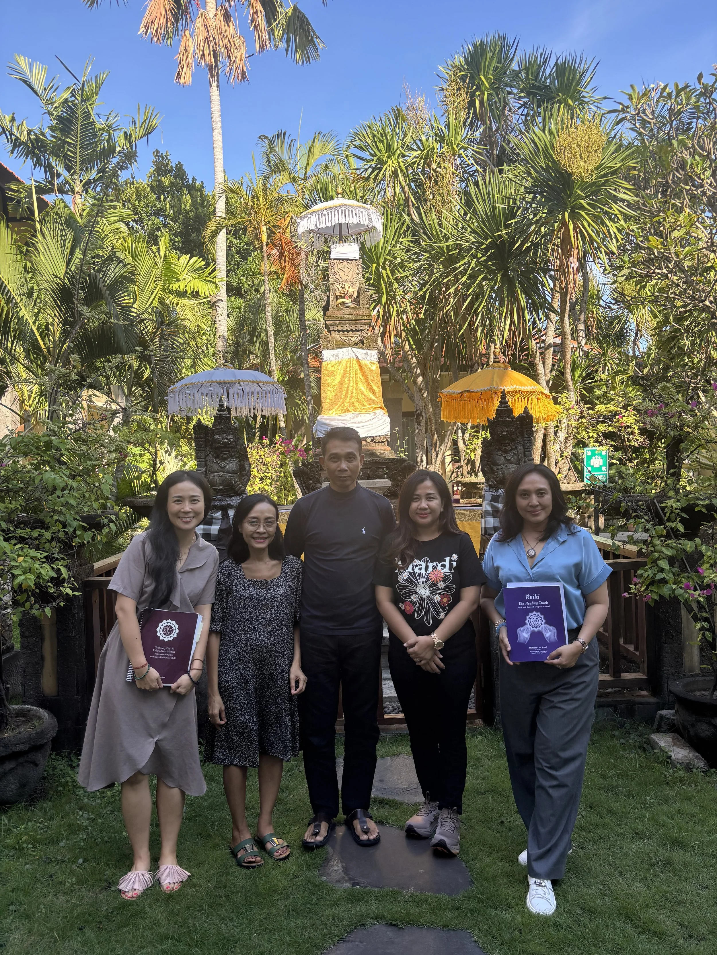 Group of five people standing outdoors in front of a small shrine with tropical plants and statues, people holding folders, bright sunny day, clear blue sky.