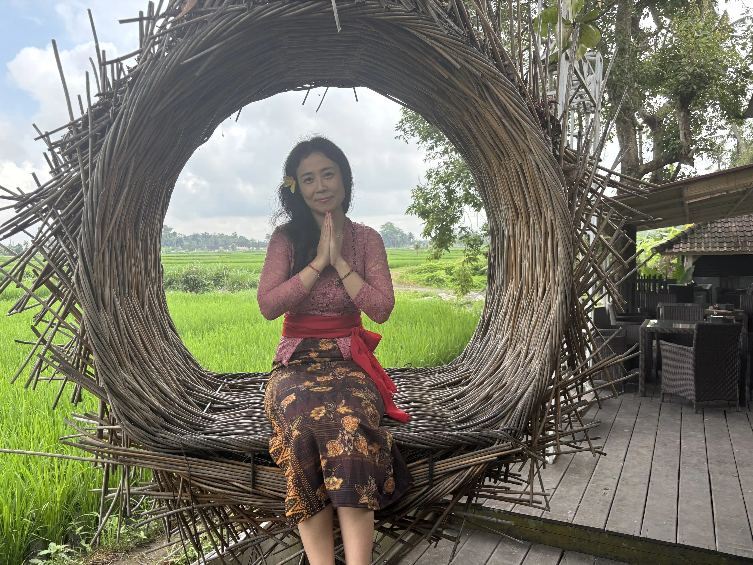 sarah lee in traditional clothing sitting inside a large, woven, circular wicker chair outside near rice fields.