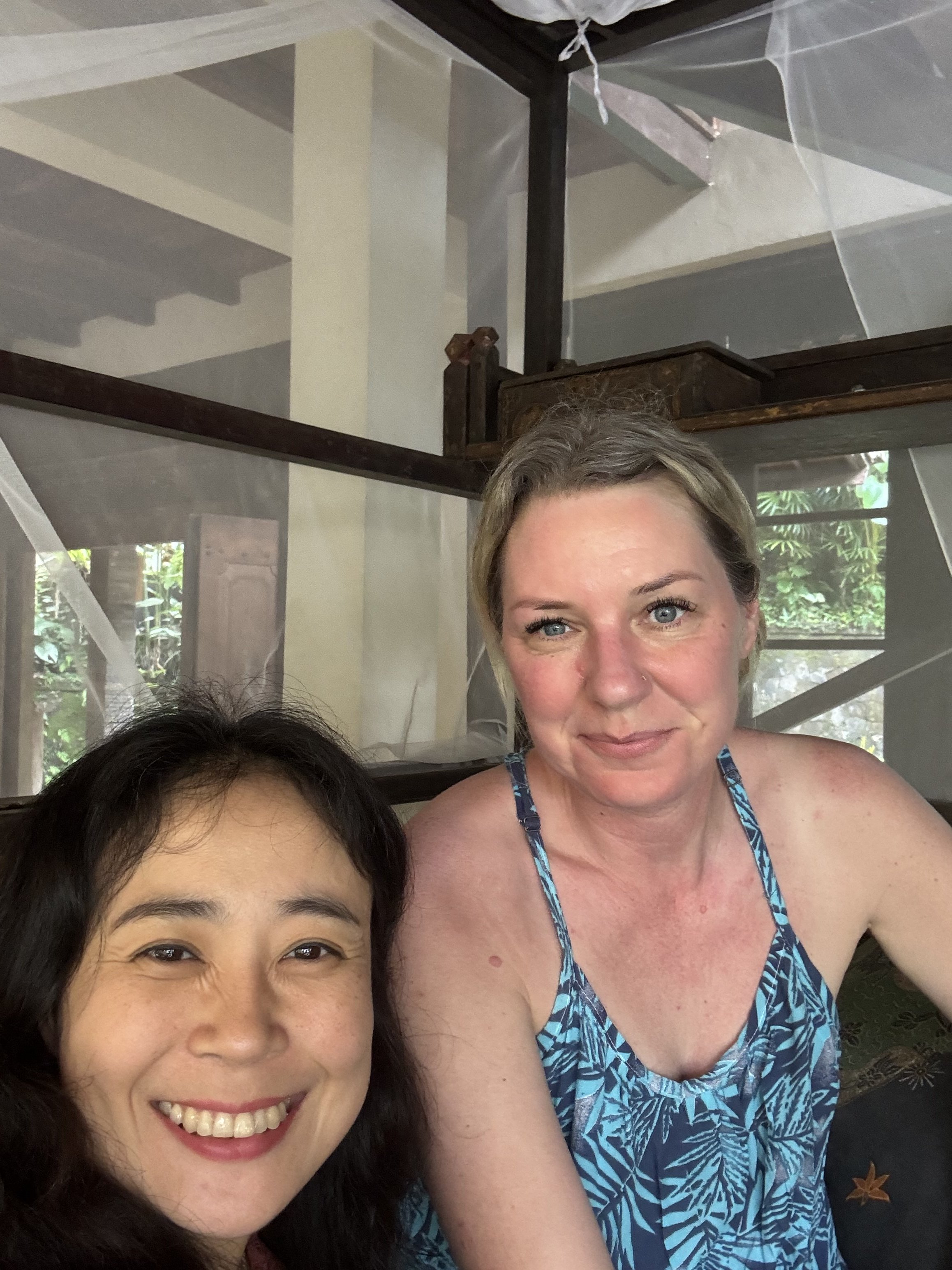 A woman with blonde hair and blue eyes wearing a blue patterned dress smiling, and a woman with dark hair and Asian features smiling, both taking a selfie in a room with large windows and greenery outside.