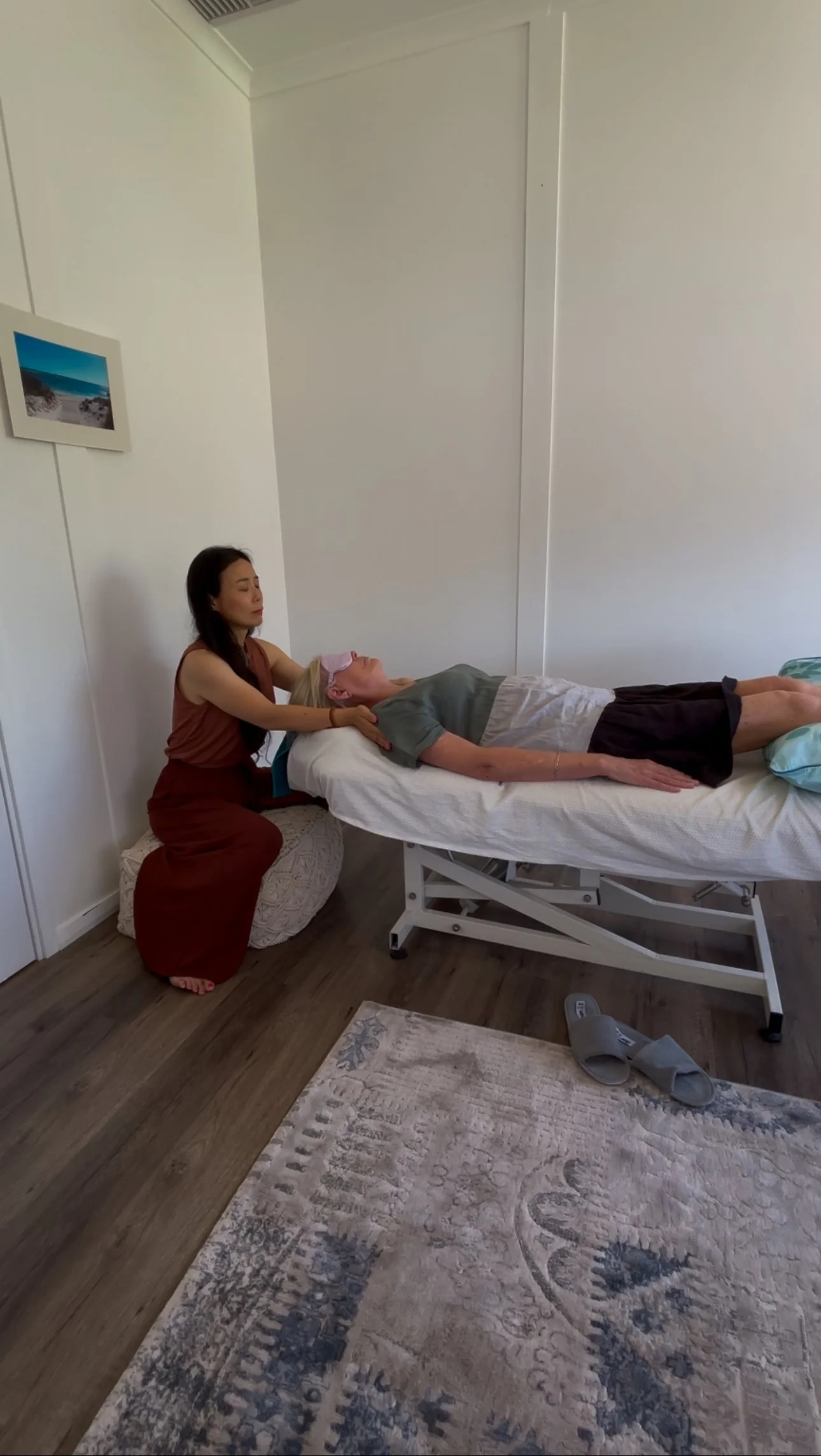 A woman lying on a medical examination table with an eye mask over her eyes, being attended to by another woman kneeling beside her in a clinical or therapy room with wooden flooring, a patterned rug, and white walls.