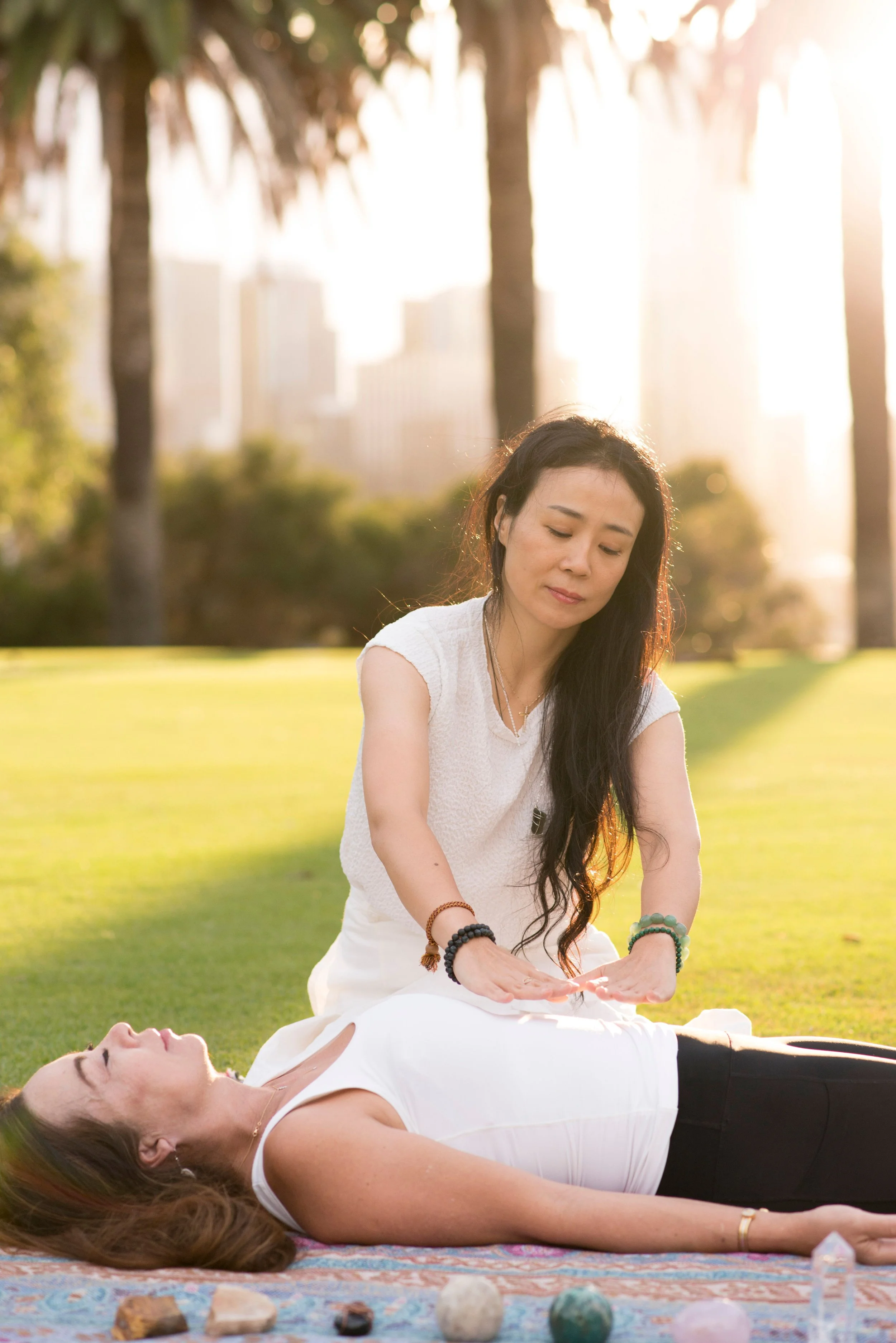 A woman lying on her back on a blanket in a park, her eyes closed and relaxed. Another woman sitting beside her, practicing Reiki or giving a healing touch, with her hands over the lying woman's stomach or chest. The scene is illuminated by sunlight, with trees and city buildings in the background.