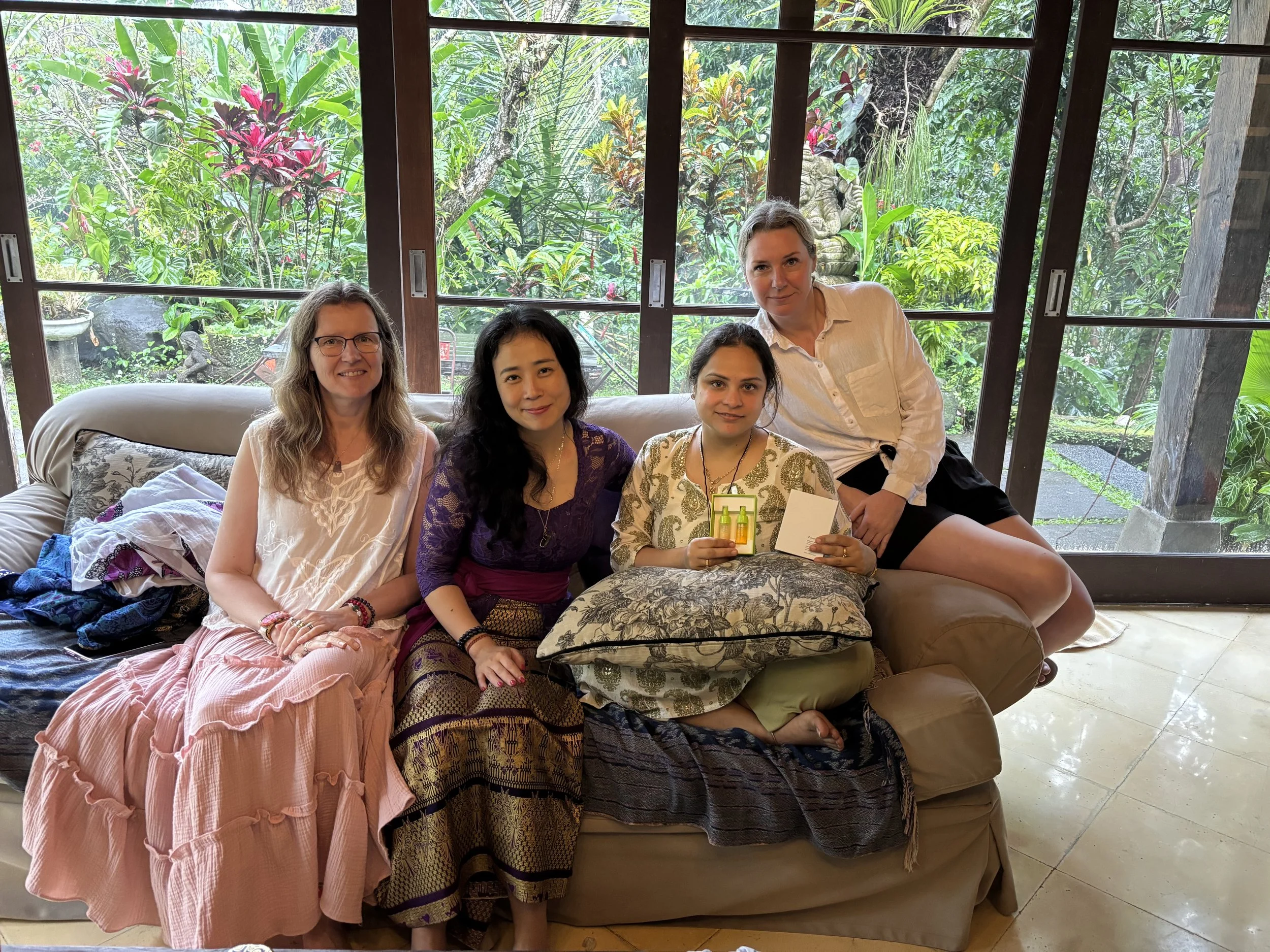 Four women sitting together on a sofa in a room with large glass windows showing a lush outdoor garden, some of them holding small bottles, smiling at the camera.