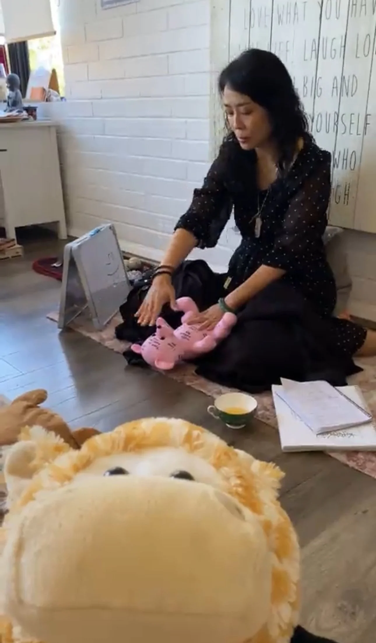 Woman sitting on the floor with a laptop, surrounded by notebooks, a cup of tea, and plush toys, in a cozy room with white brick walls and a handwritten poster.