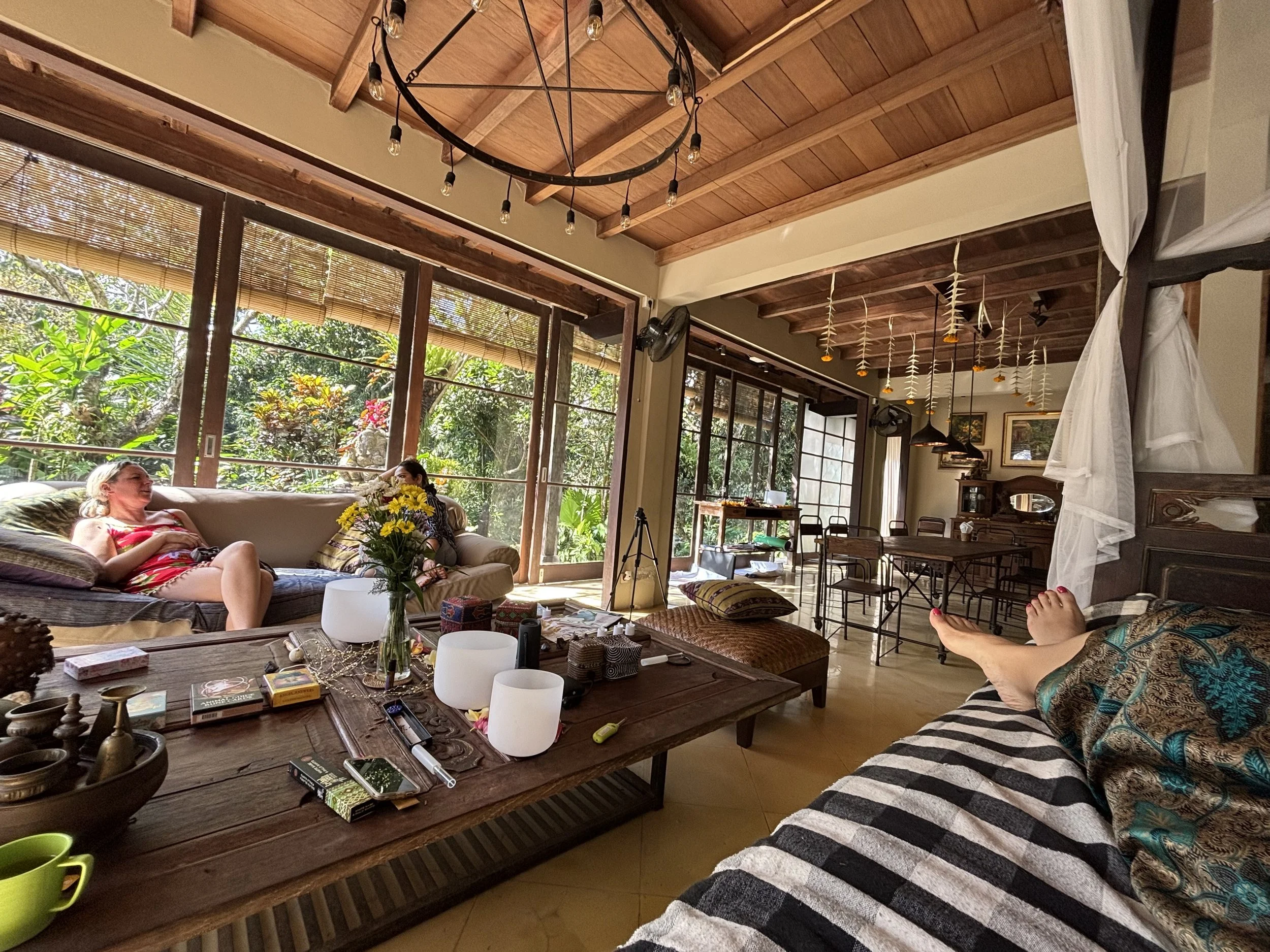 Living room with wooden ceiling, large windows, and natural light, decorated with tropical plants, a chandelier, and hanging décor, with people relaxing on a sofa.