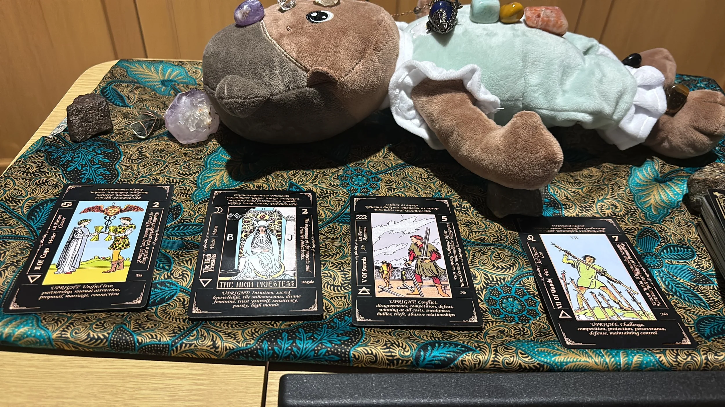 A table with a black cloth featuring blue and gold floral patterns, holding tarot cards, crystals, and a stuffed animal with rocks and gemstones on its back.