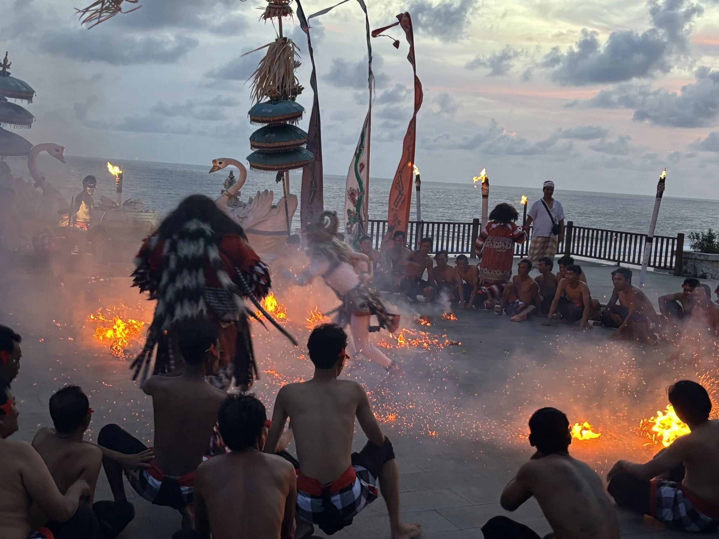 A group of people gathered on a beach near the ocean during sunset, participating in a traditional ceremony with flames, tiki torches, and flags, with some individuals seated and others standing.