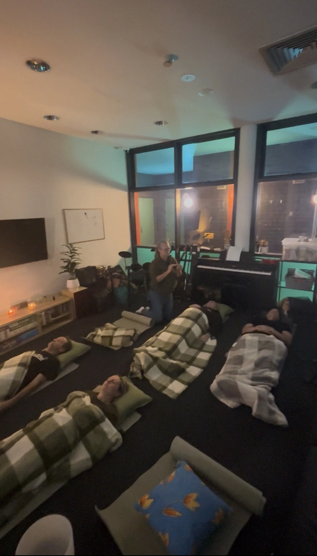 People are lying on mats with blankets in a dimly lit room, possibly participating in a relaxation or meditation session, with a woman standing and holding a phone or camera.