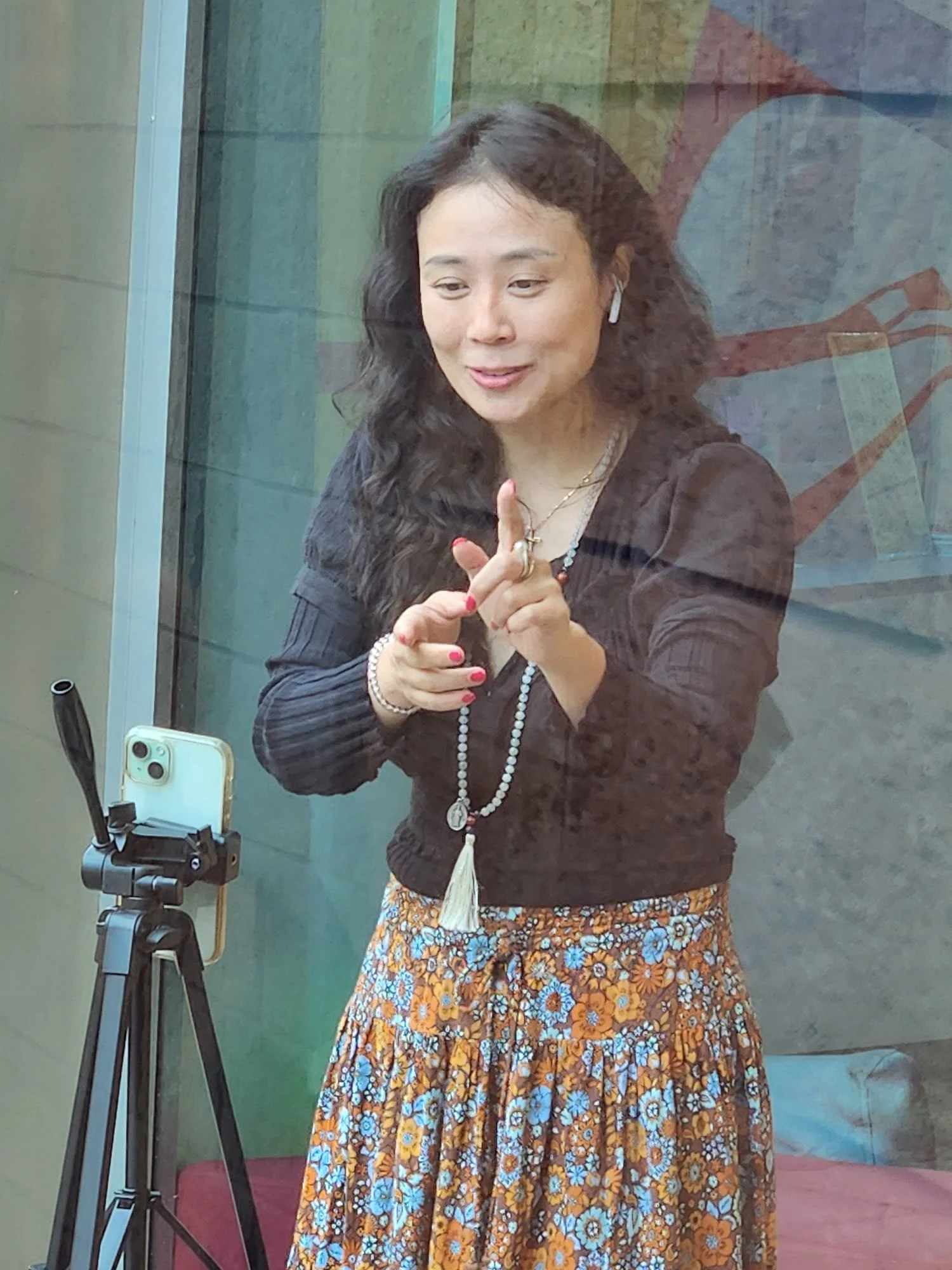 A woman with wavy black hair wearing a black blazer, colorful floral skirt, layered necklaces, and white wireless earbuds, standing behind a glass window taking a selfie with a smartphone on a tripod.