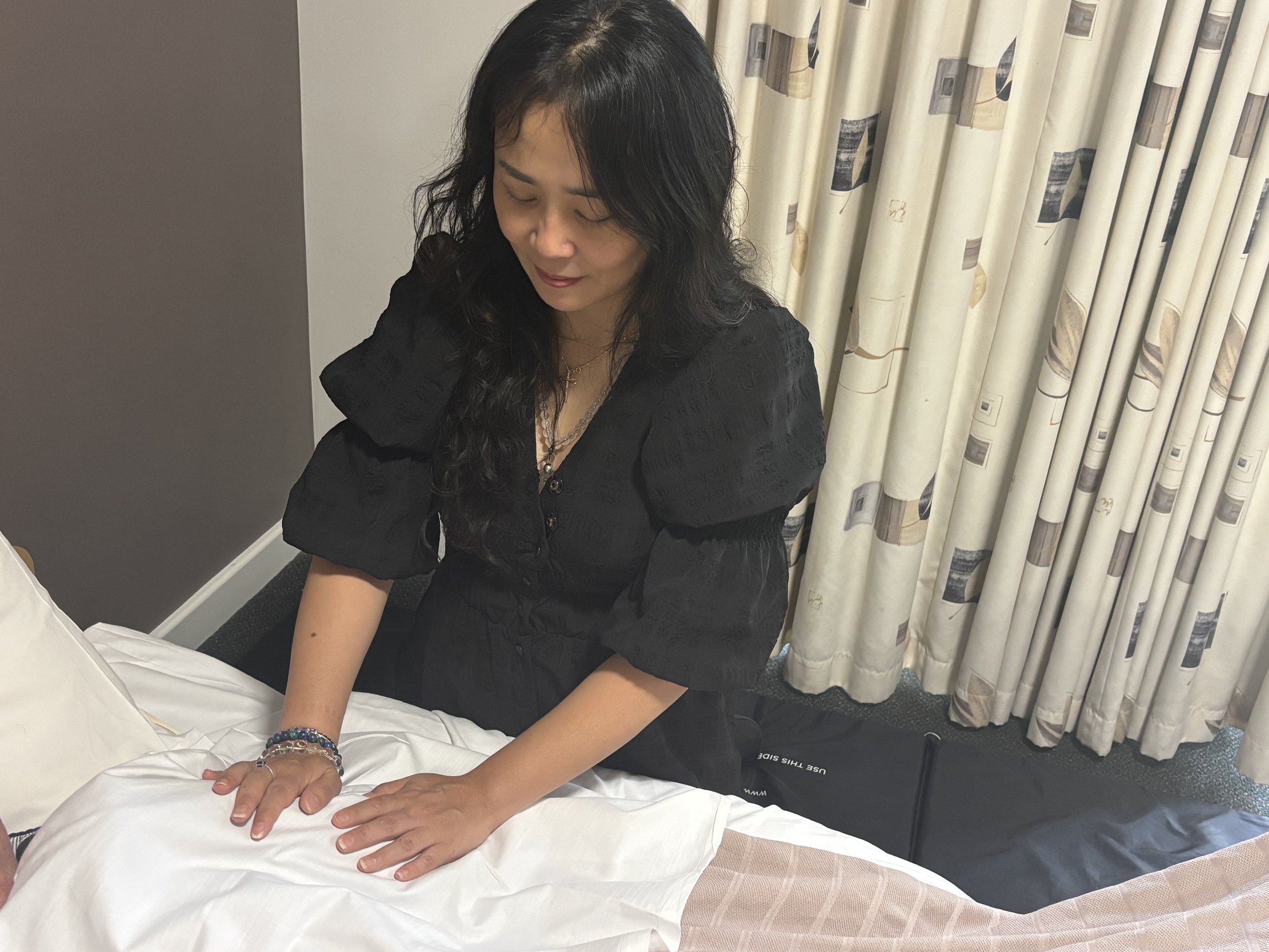 A woman with long black hair, wearing a black top and jewelry, is sitting on a bed with white sheets, her eyes closed, and her hands resting on a pillow. Behind her are patterned curtains and a gray wall.