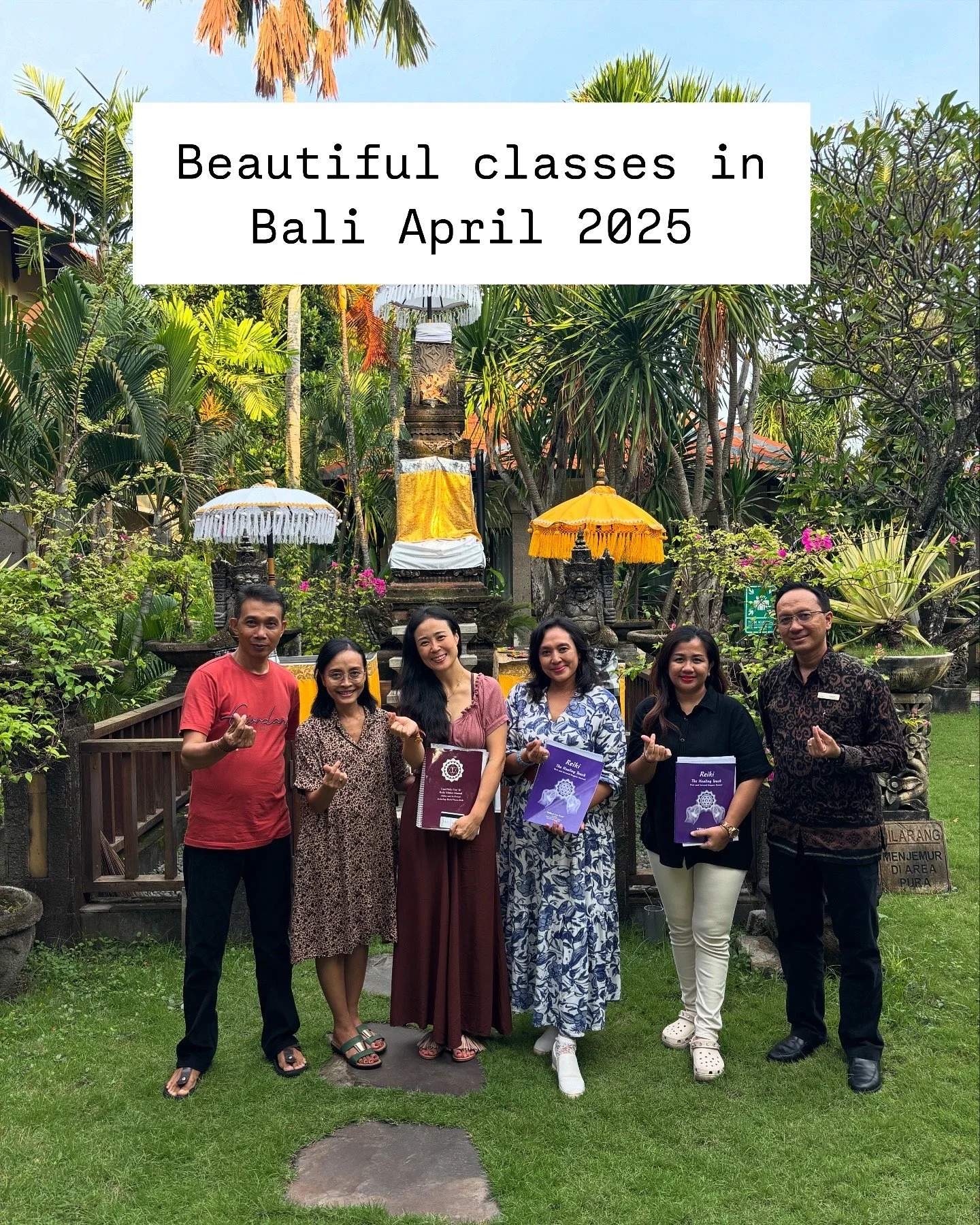 Group of six people standing outdoors in a lush, tropical garden with statues and parasols behind them. They are dressed in semi-formal attire and holding books, smiling at the camera. A white banner with black text overlays the top of the image, rea