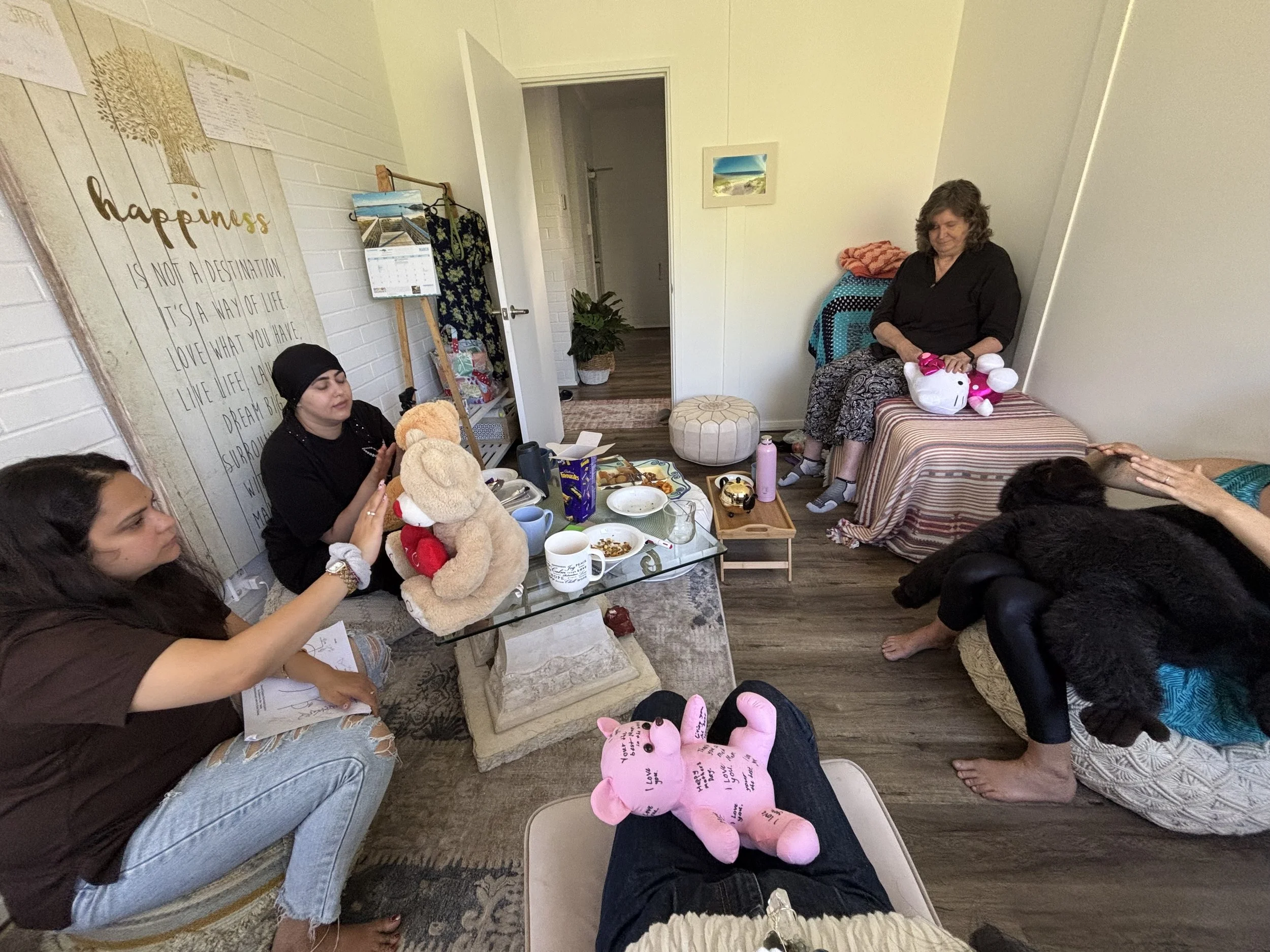A group of people gathered in a cozy indoor space with a table set for food and drinks, some sitting and others standing or sitting on a bench. The room is decorated with framed pictures, a poster that reads 'happiness is not a destination, it’s a way of life,' and a potted plant. There are gifts, plush toys, and miscellaneous items on the table and around the room.