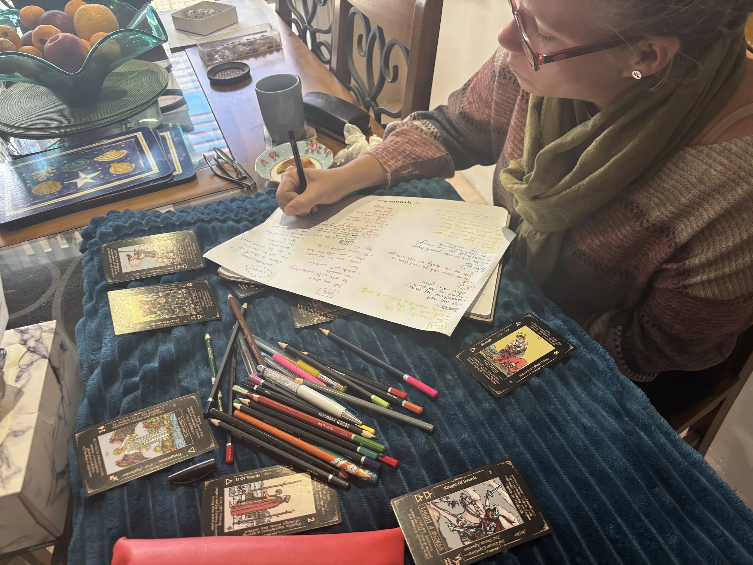 A woman sitting at a table with Tarot cards spread out, along with colored pens, glasses, a mug, and a bowl of fruit on the table.