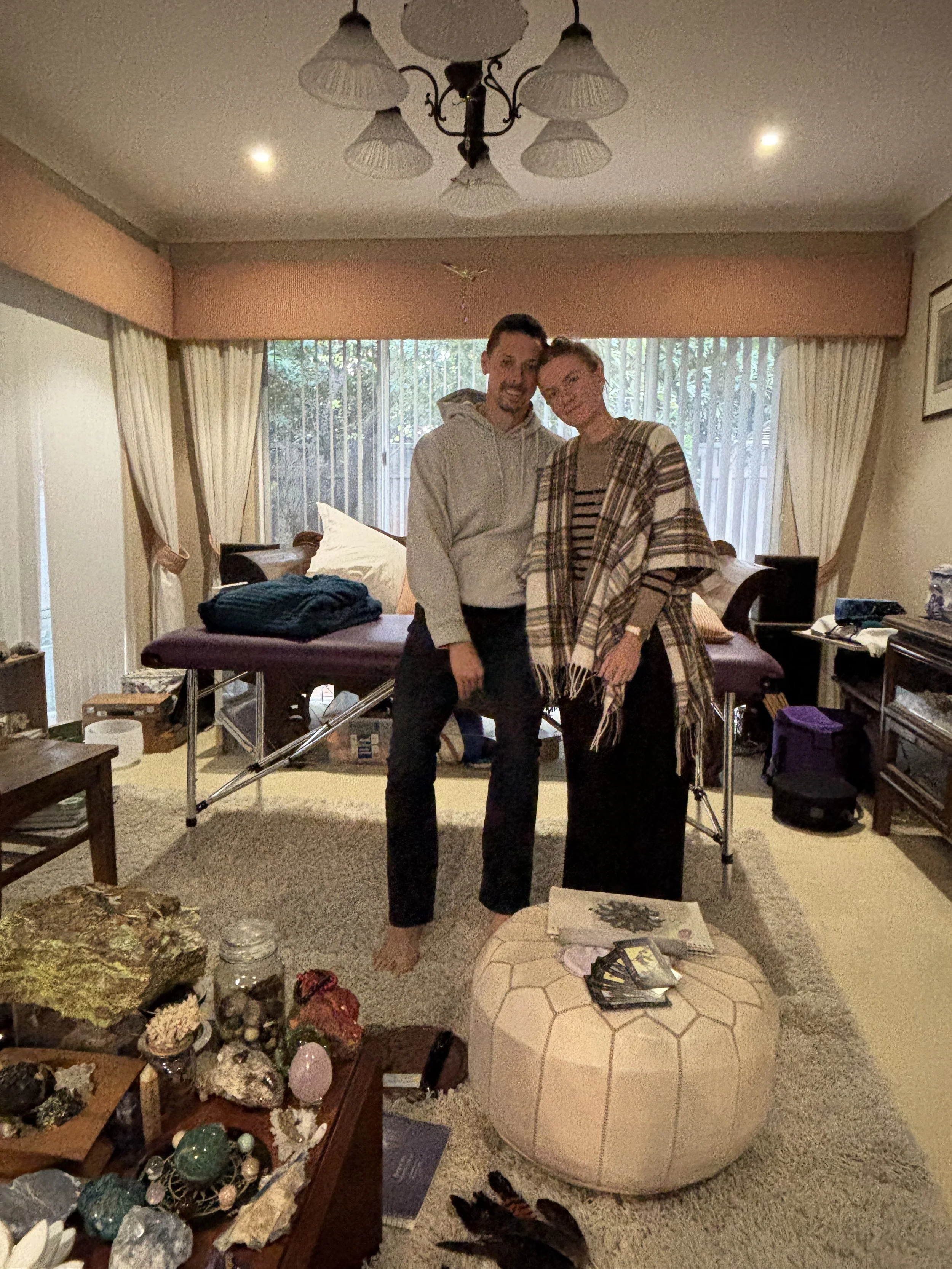 Two people stand close together in a living room, smiling at the camera. Behind them is a large window with vertical blinds, and an organization table with rocks, jewelry, and miscellaneous items. The room has a beige carpet and a pool table covered 