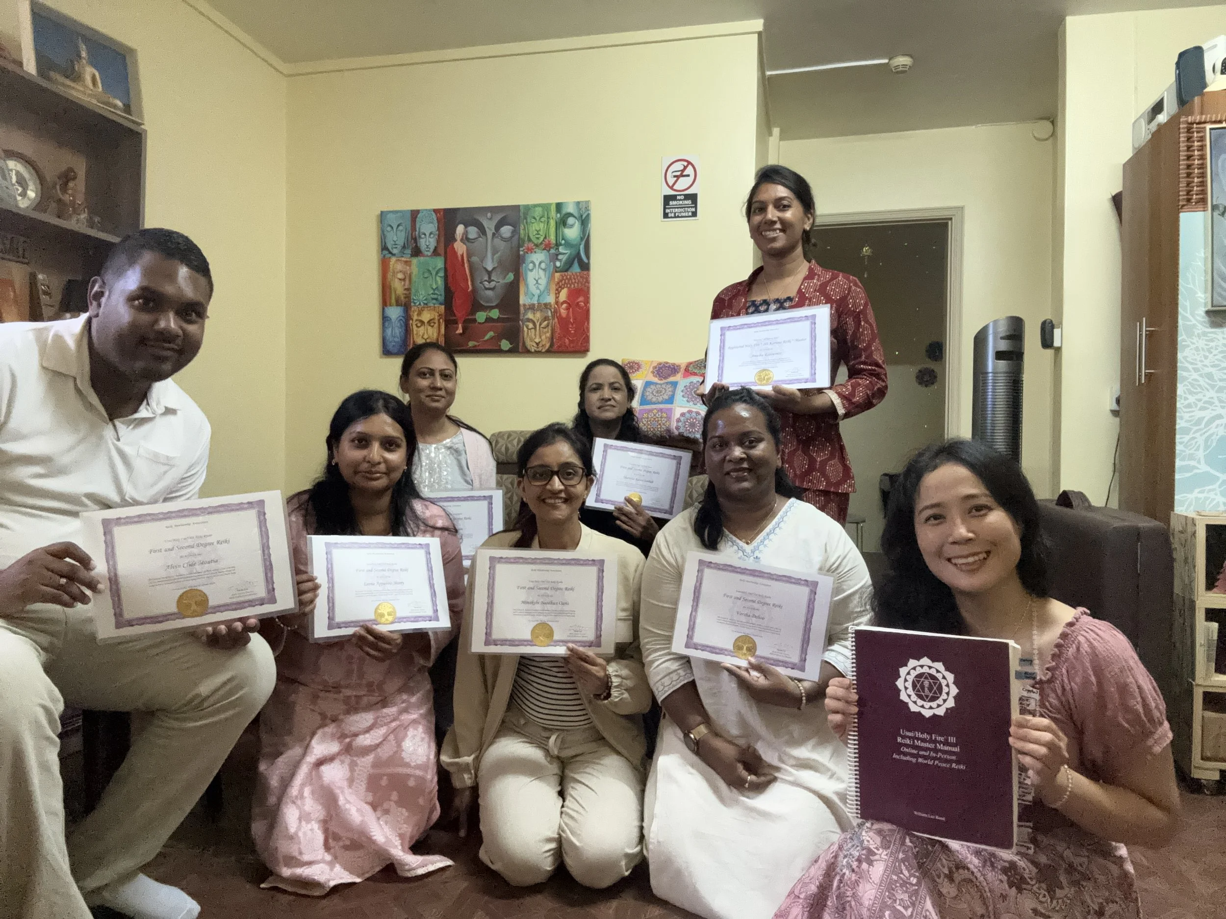 Group of women and a man celebrating a graduation or certification ceremony in a living room, holding certificates and a book, with colorful wall art behind them.