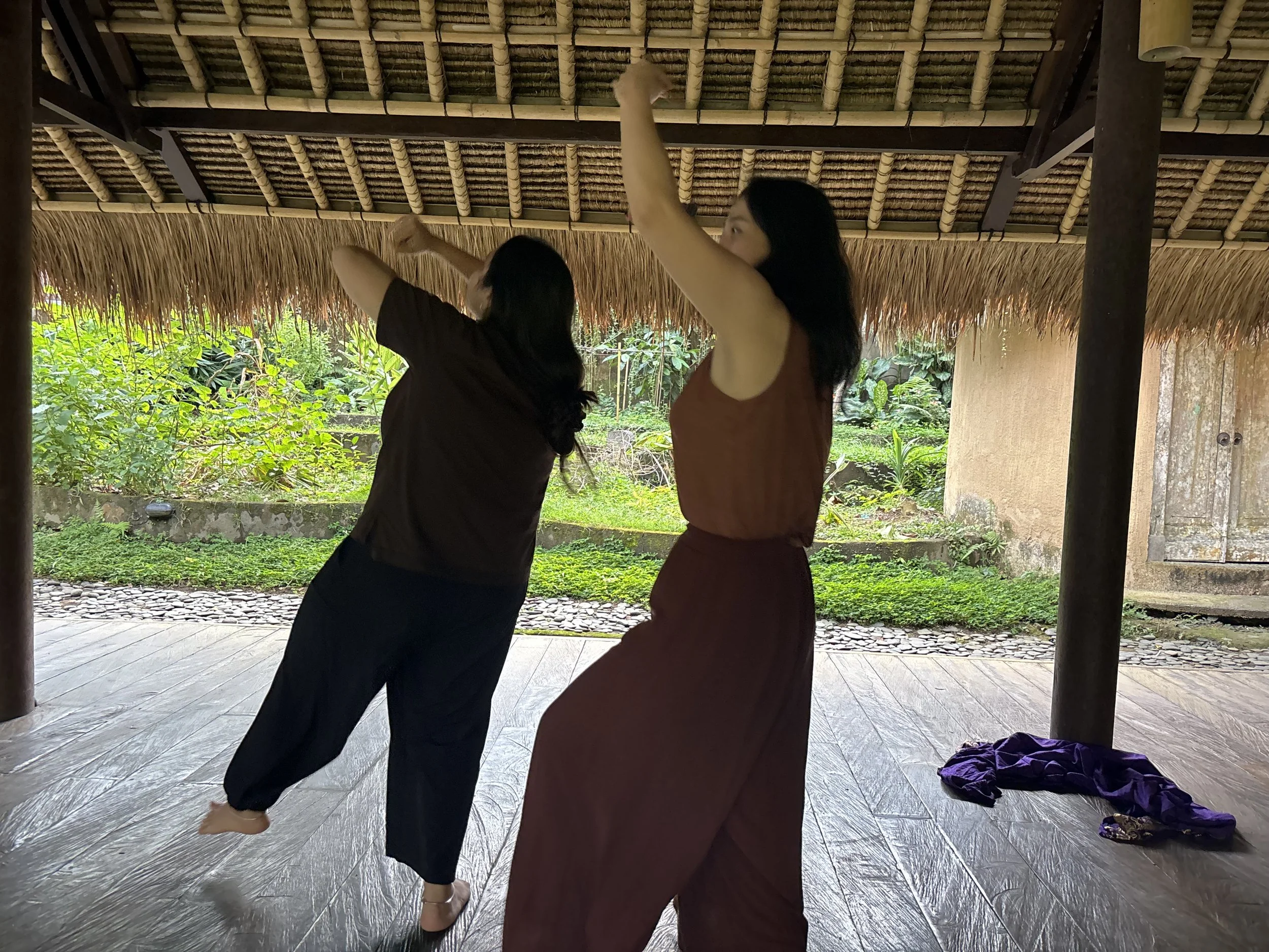 Two women dancing or practicing dance indoors with a thatched roof ceiling, green garden visible outside, and some clothing on the wooden floor.