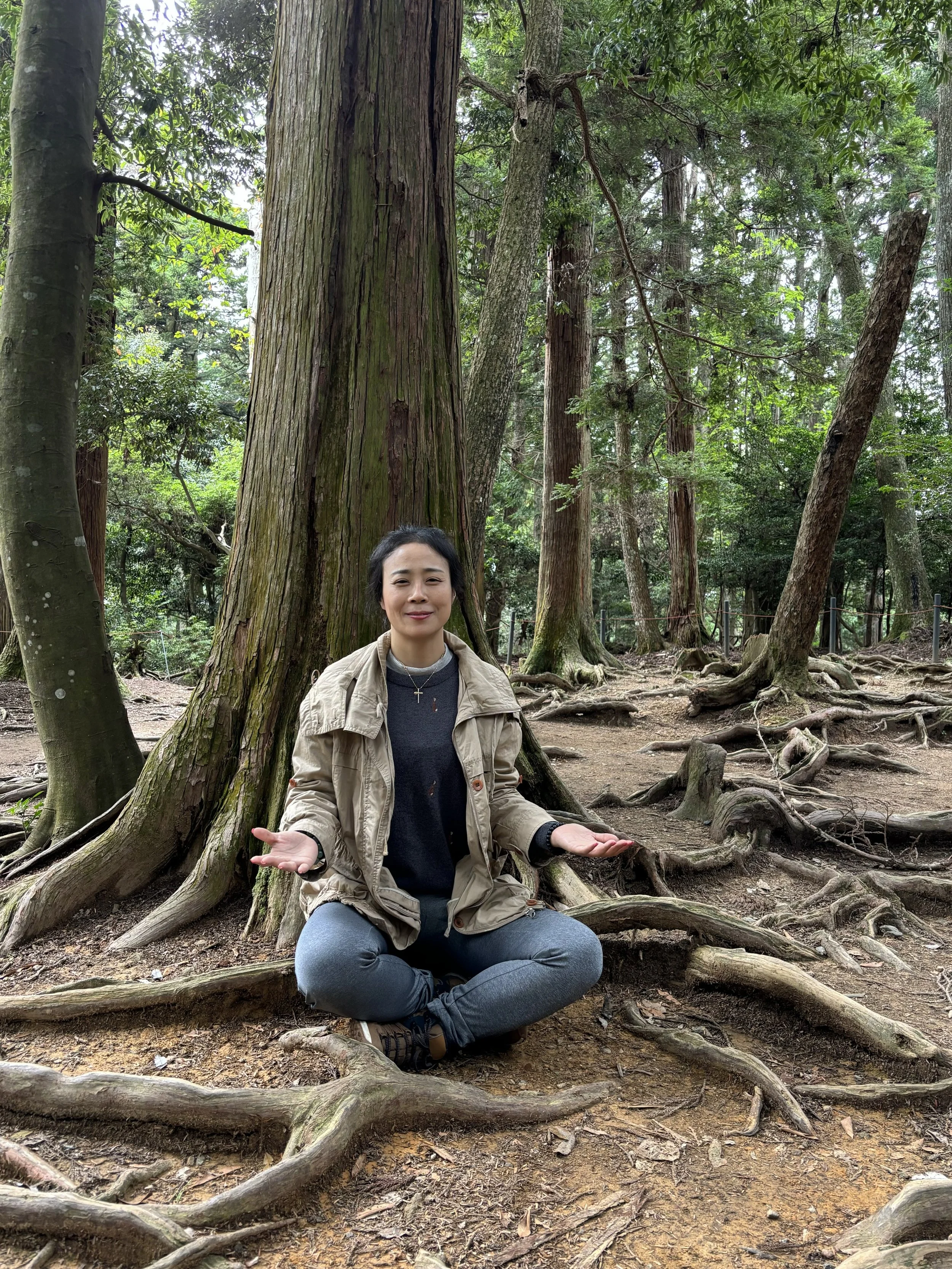 A woman sitting cross-legged on the forest ground with roots and large trees around her, making a shrugging gesture with both hands and smiling.