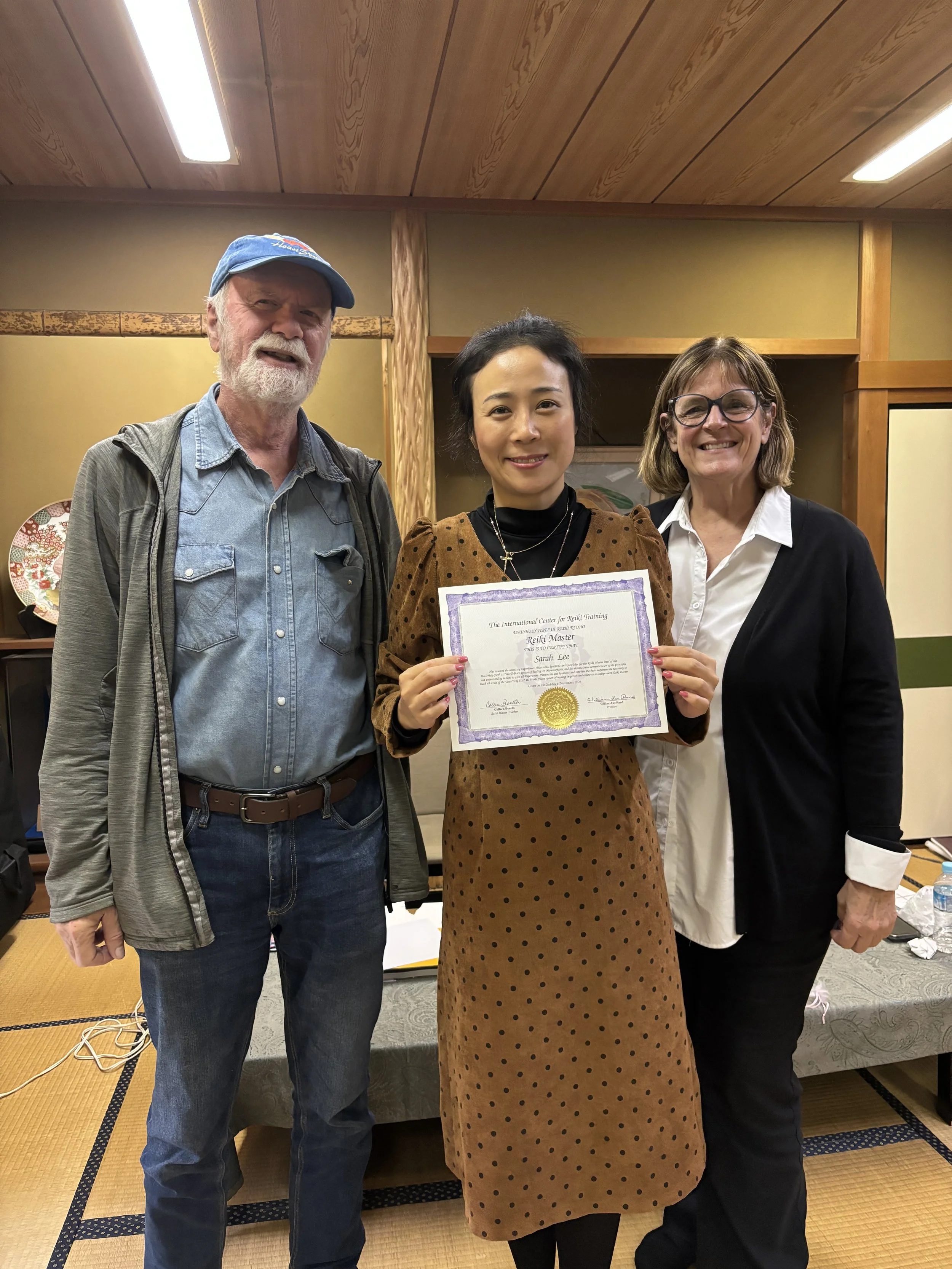 Three people standing indoors, smiling, with the woman in the middle holding a certificate. The setting appears to be a conference or ceremony room with traditional Japanese tatami mats on the floor.