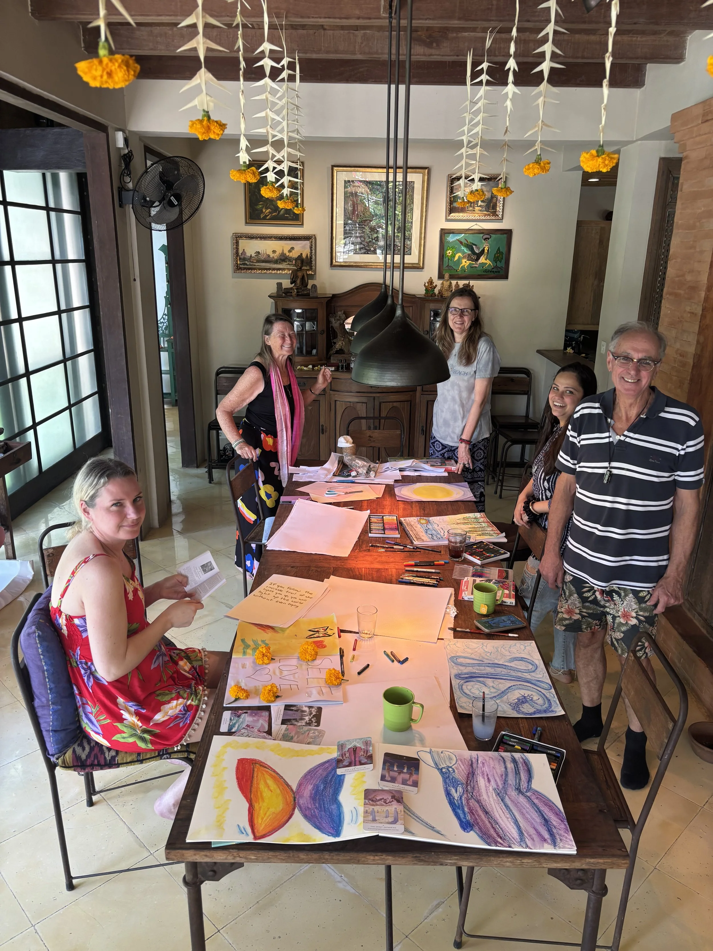 People participating in a creative art activity in a bright room with wooden ceiling beams, decorated with hanging paper stars and marigold flowers. The table is covered with colorful drawings, markers, and art supplies.